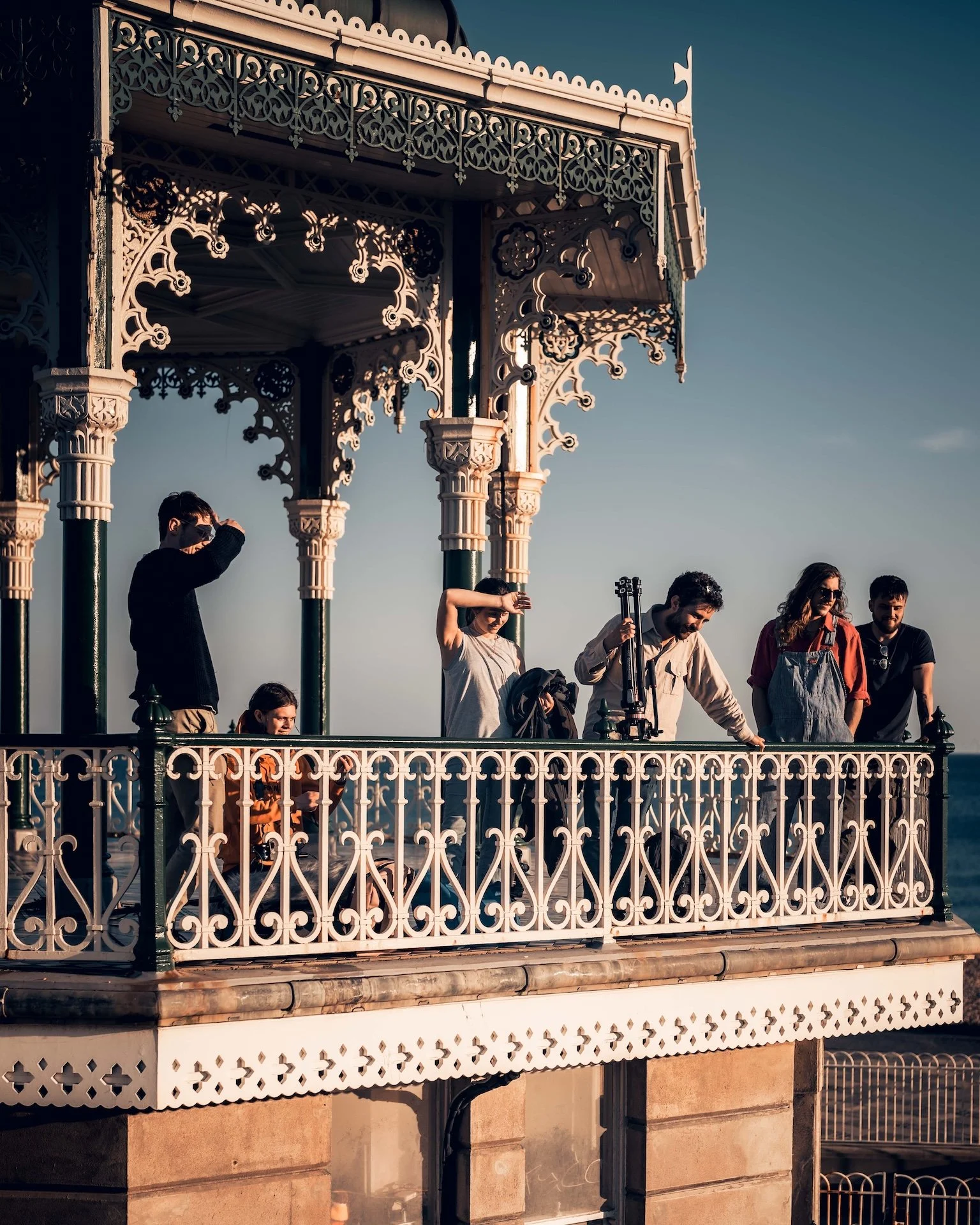 A group of people on a decorative balcony with ornate ironwork, overlooking the ocean during sunset.