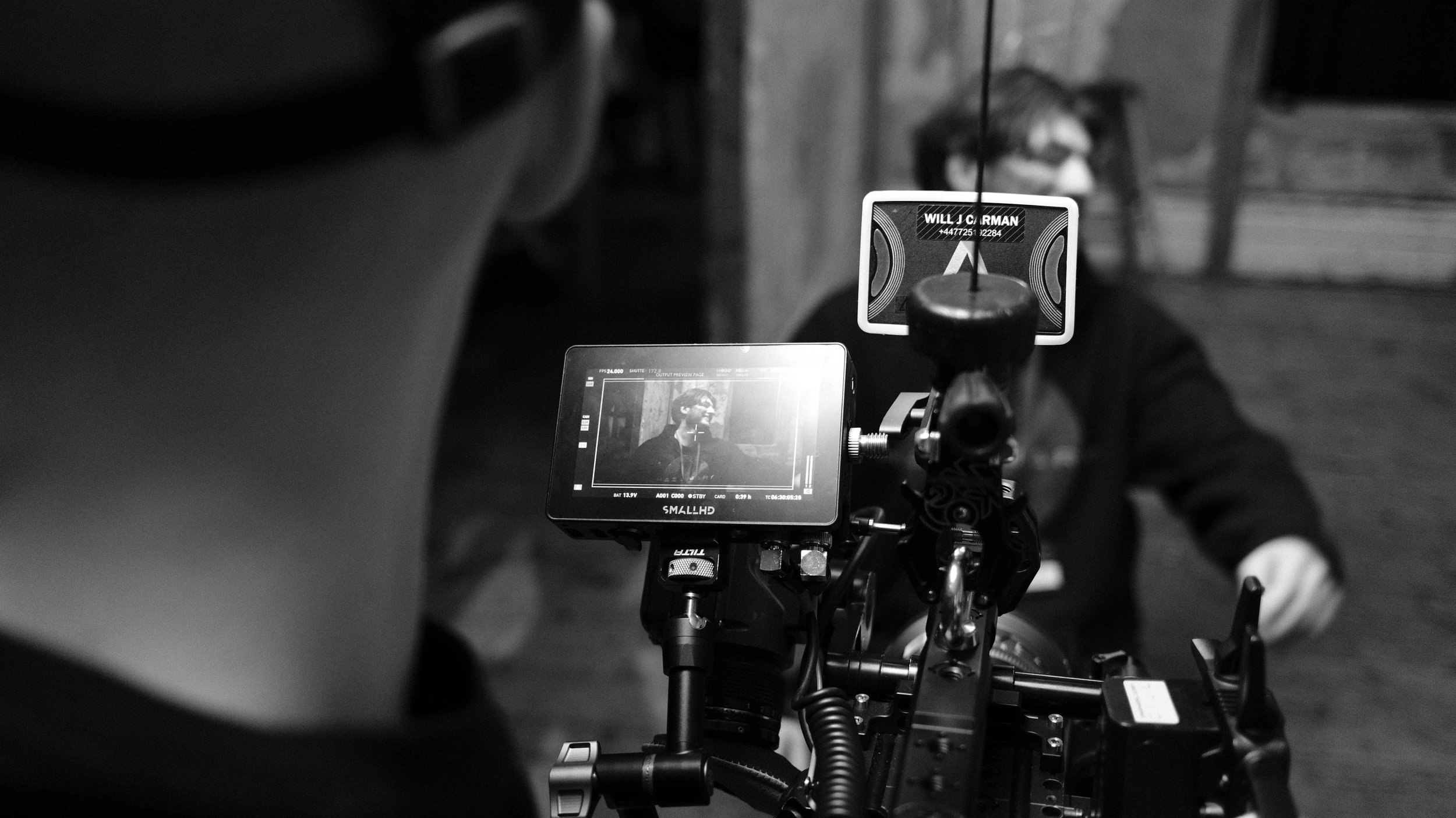 Black and white photo of a film or video set showing a camera on a tripod and a monitor displaying a person with glasses in the background.
