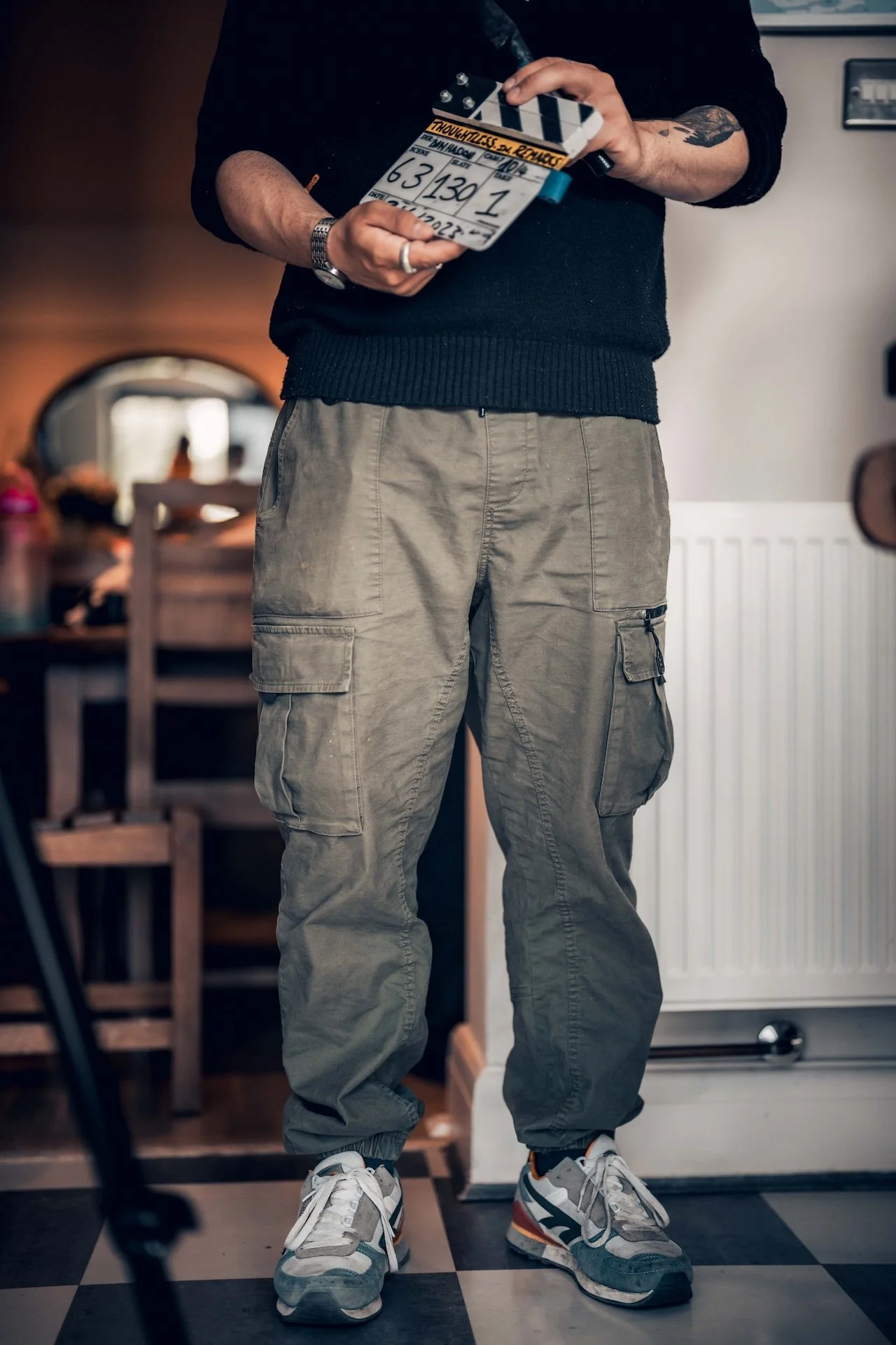 Close-up of a person holding a clapperboard in a room with wooden furniture and checkered floor tiles. The person is wearing cargo pants, sneakers, and a black long-sleeve shirt.