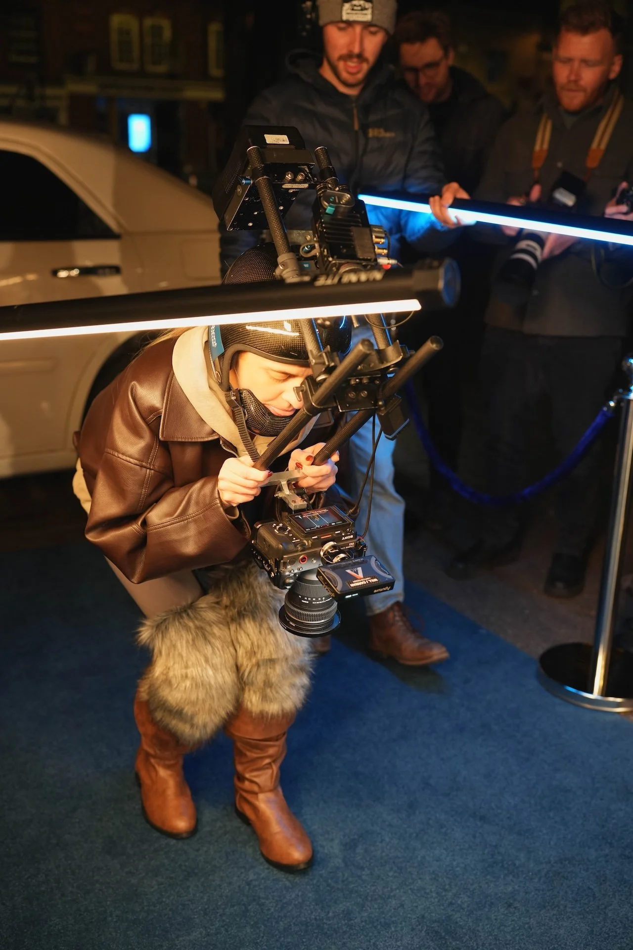 A young woman wearing a brown leather jacket, boots, and a black helmet is operating a professional film camera on a stabilizer rig, with a furry pom-pom accessory hanging from her jacket. She is inside a building, with three men in the background ex