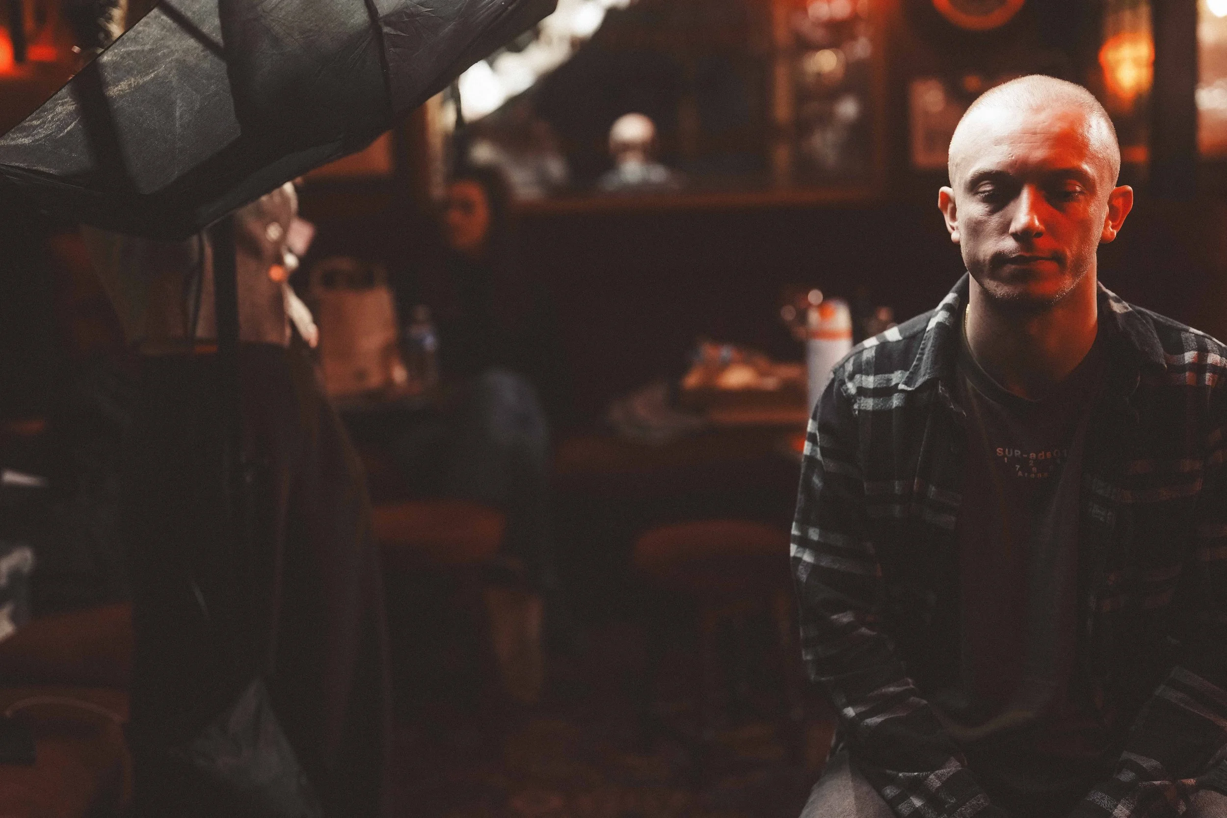 A person with a buzz cut hairstyle wearing a black and white plaid shirt in a dimly lit bar or pub setting with a red hue, sitting on a barstool with another person partially visible in the foreground.