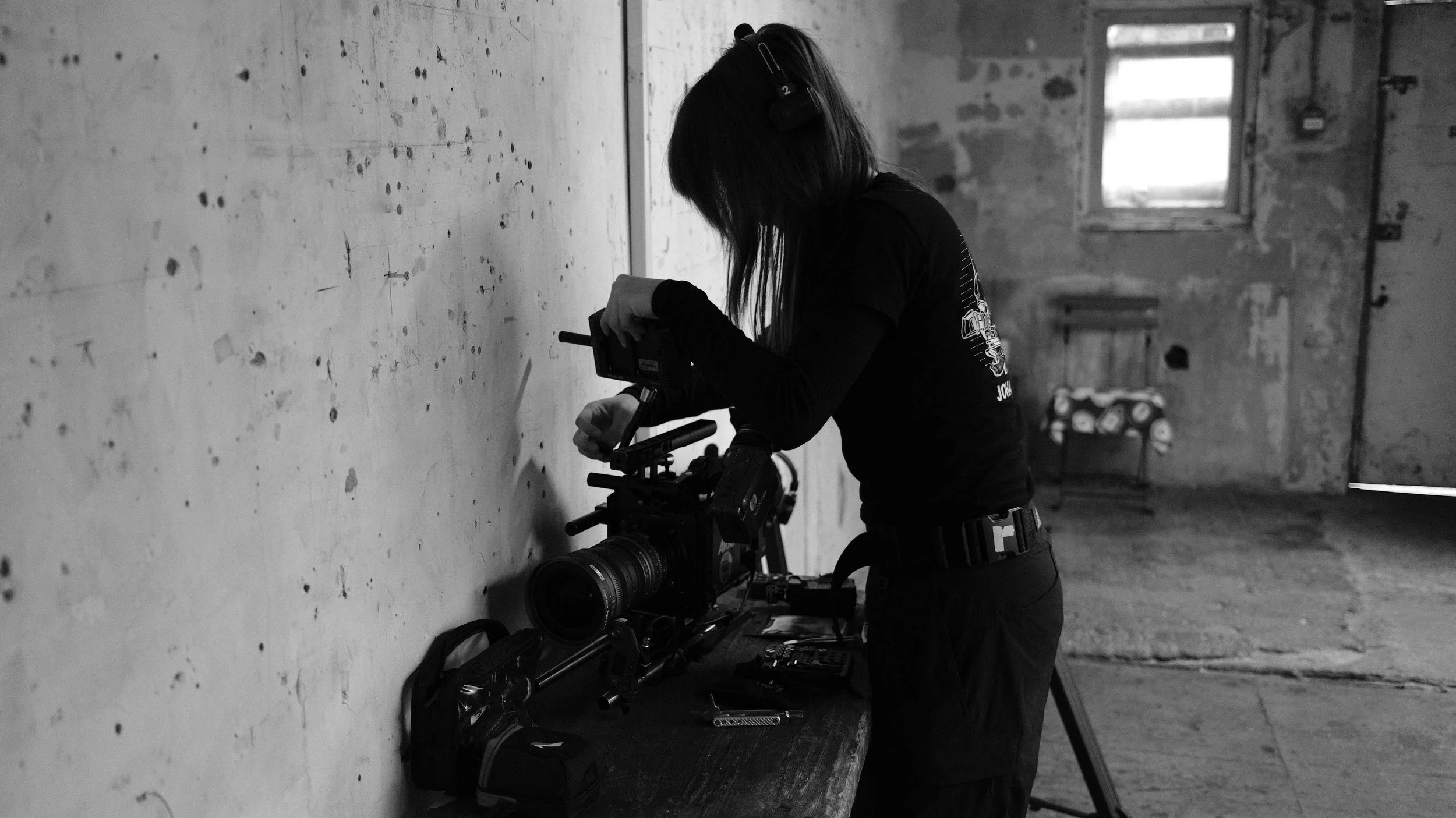 A woman setting up a professional video camera on a tripod in an industrial-style room with distressed walls and a small window.