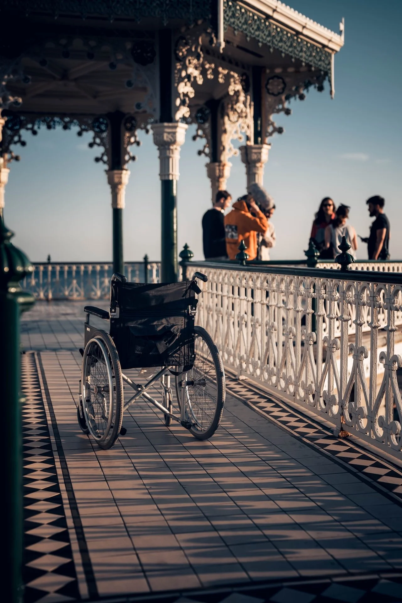 Empty wheelchair parked on a black-and-white tiled promenade at a seaside pier, with people socializing under a cabana in the background during evening.