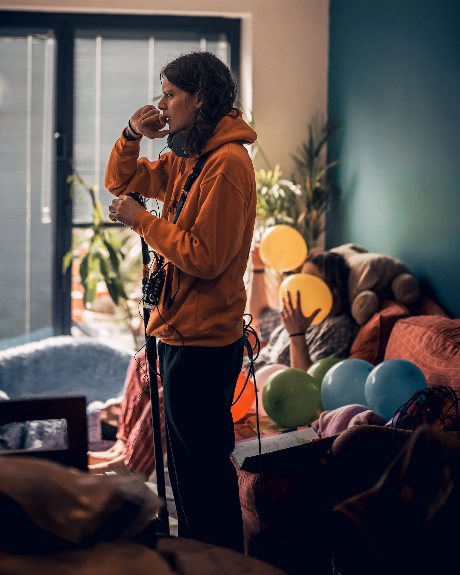 A young man with long hair and a brown hoodie, wearing headphones, is standing near a window in a room decorated with colorful balloons. Two women are sitting on a couch in the background, one holding balloons and the other resting her head on the co