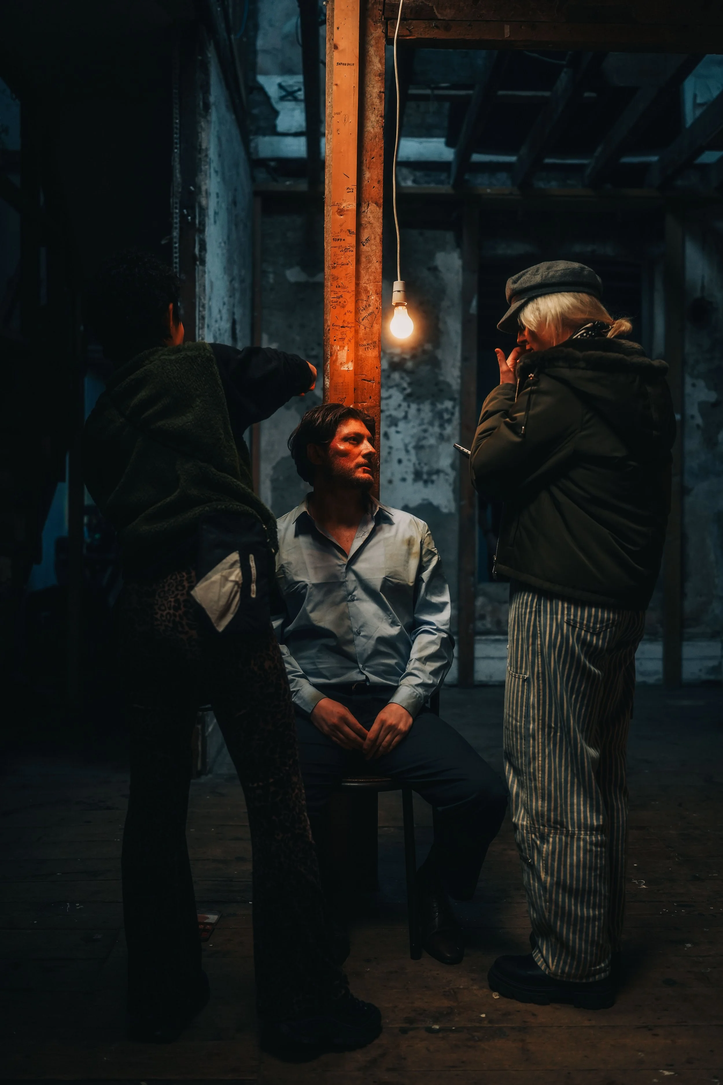 Three people conversing in an industrial setting, one person sitting on a chair, two standing, with a single exposed light bulb hanging from the ceiling, and walls with peeling paint and exposed wooden beams.