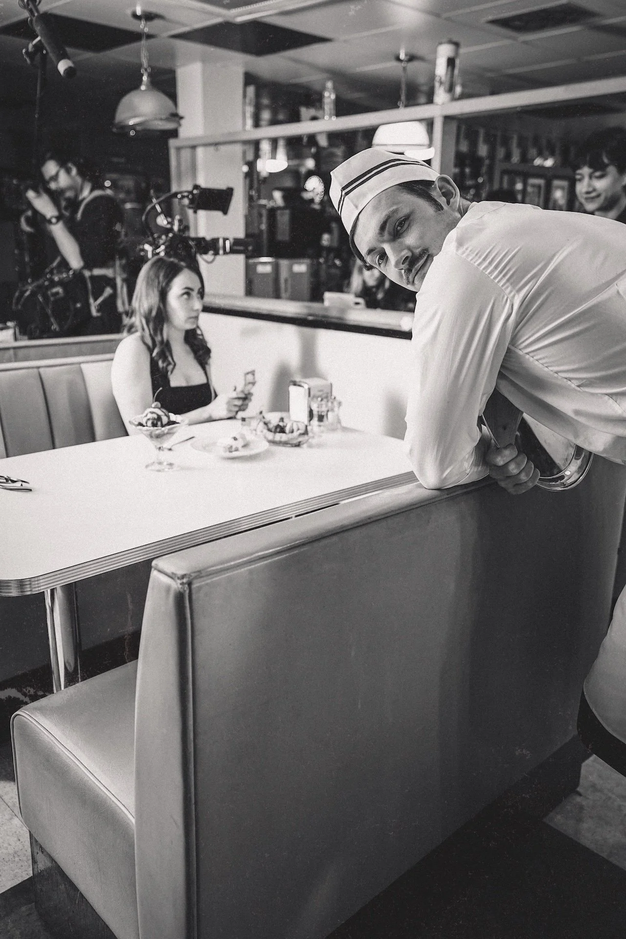 Black and white photo of a male waiter leaning on a table in a diner, with a woman sitting at the table looking at her phone, and a person with a bicycle in the background.