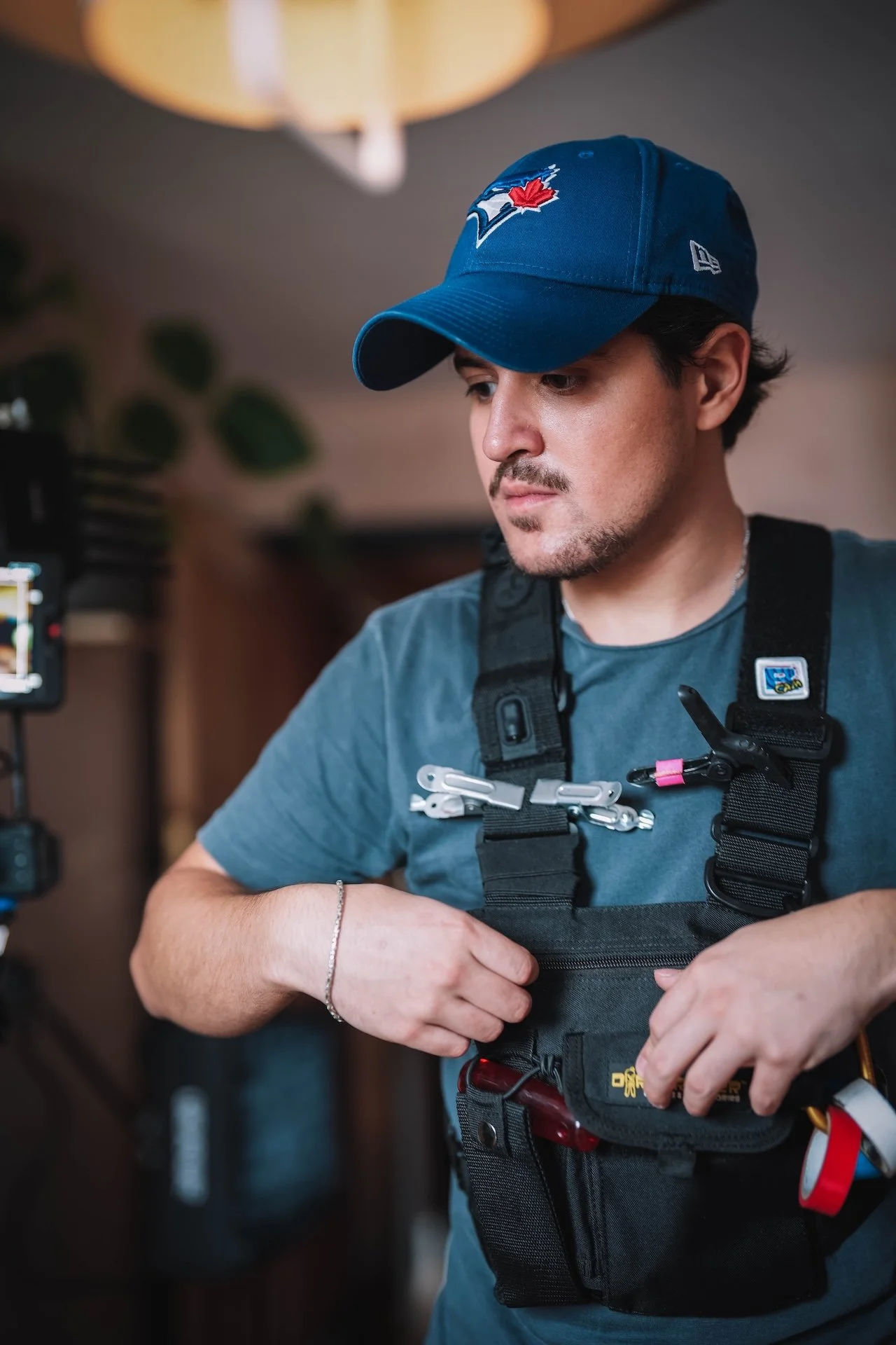 A man wearing a blue baseball cap with a Toronto Blue Jays logo, a dark gray t-shirt, and a black utility vest with various tools and clips. He is focused on adjusting his vest indoors.