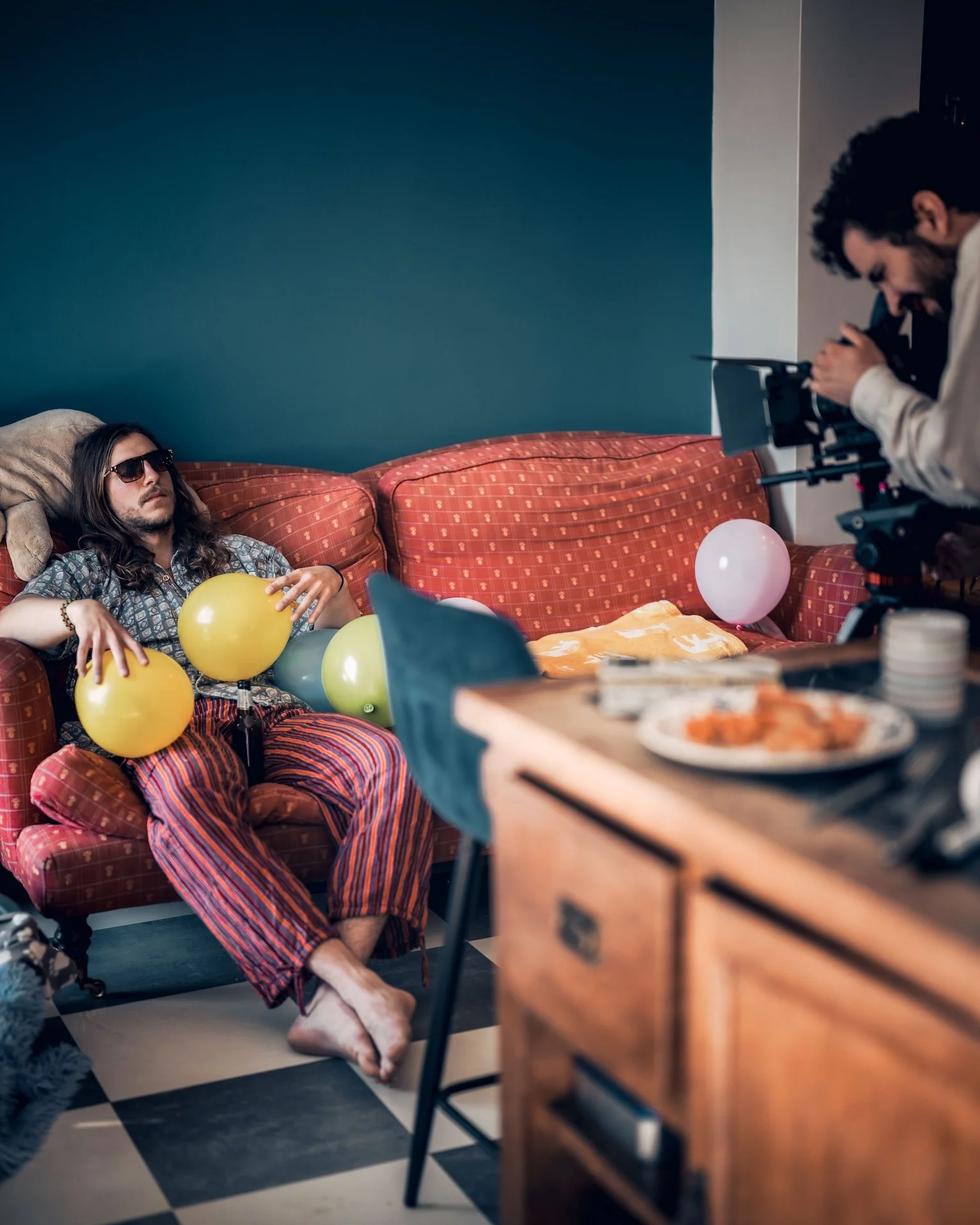 A young man with long hair and sunglasses sitting on a red couch at a party, surrounded by balloons, wearing striped pants and a patterned shirt, while a photographer takes his picture.