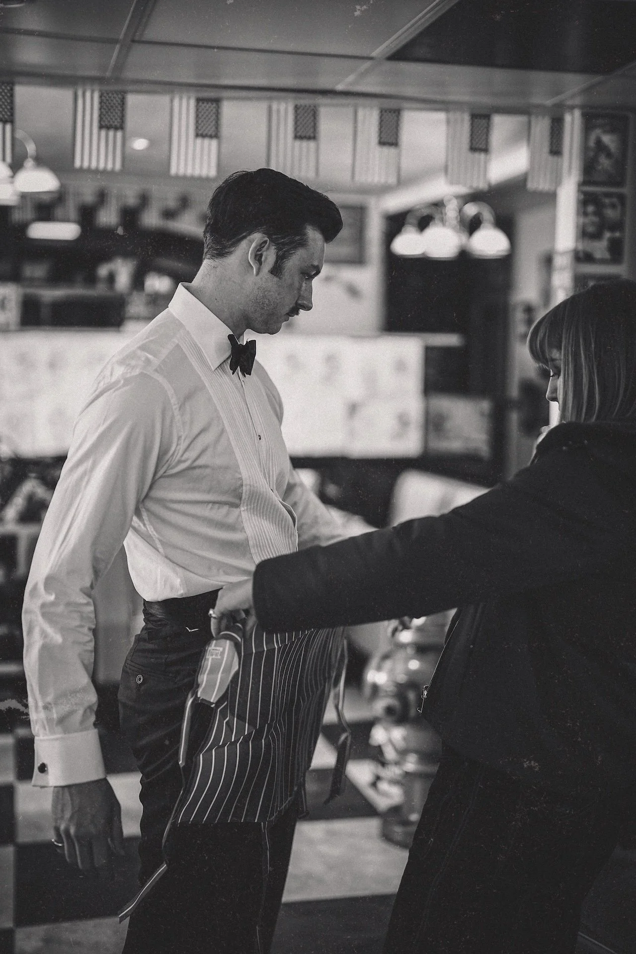 A male waitress in a white shirt, black bow tie, and striped apron standing at a restaurant counter, talking to a female customer in a dark jacket with long hair.