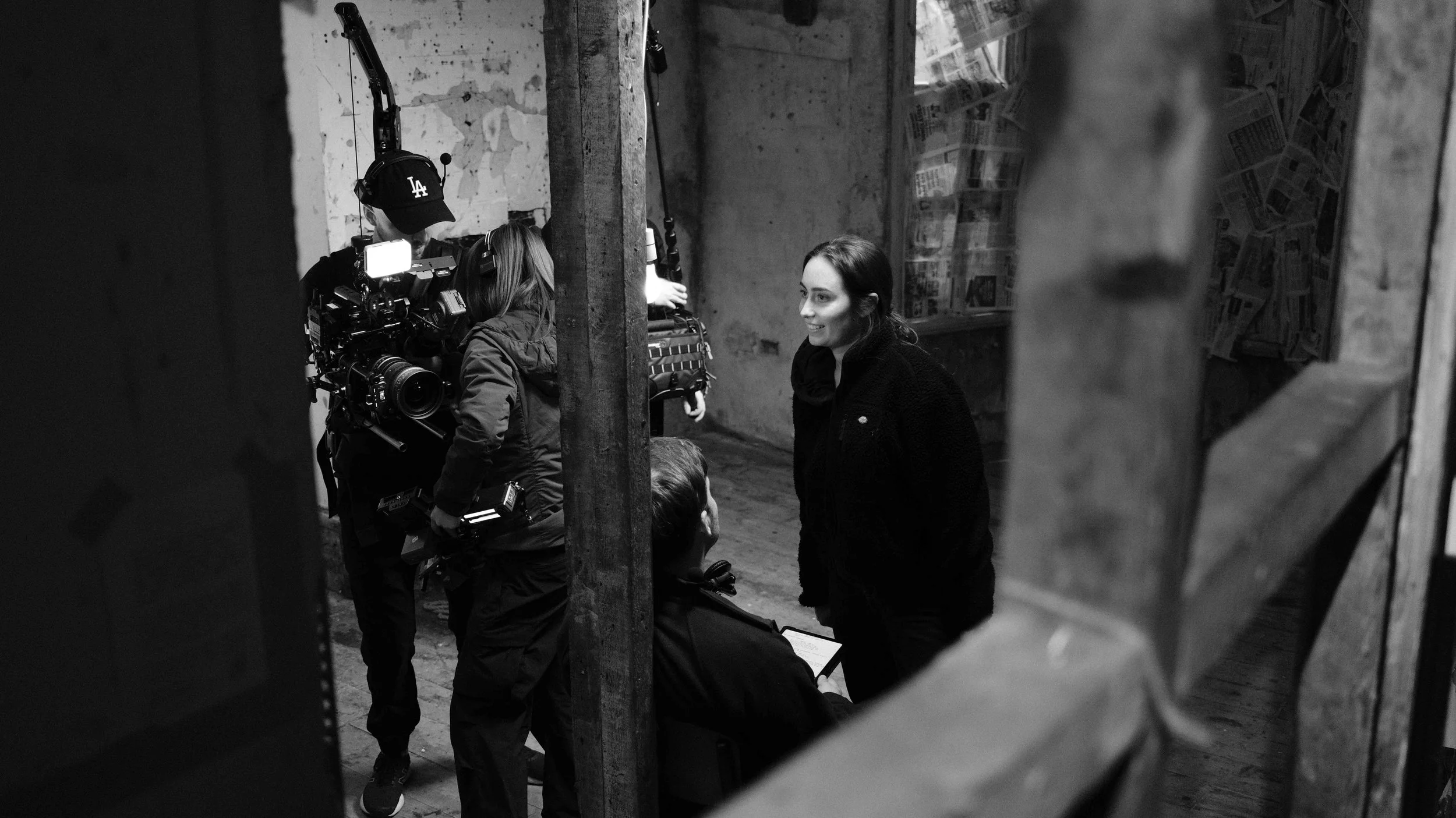 A woman is smiling during a film shoot with a crew including a camera operator and a director in an industrial setting.