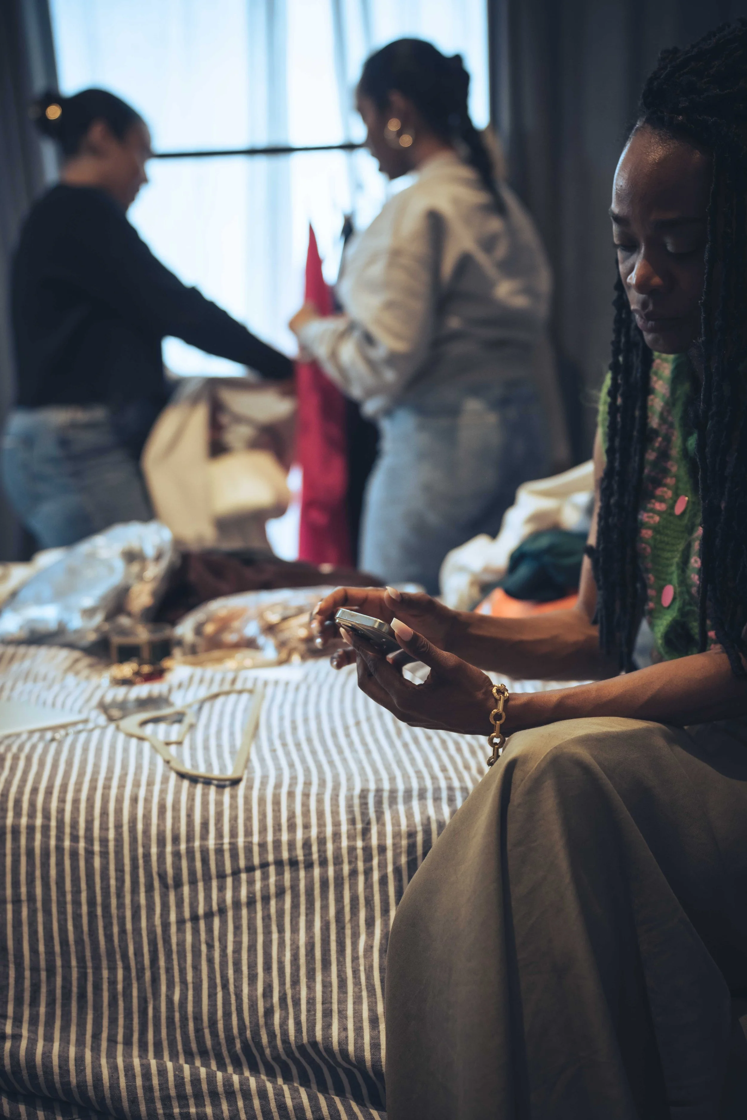 Three women with braided hair in a room, one in the foreground looking at her phone, and two in the background, one handling items on a table covered with a striped cloth.