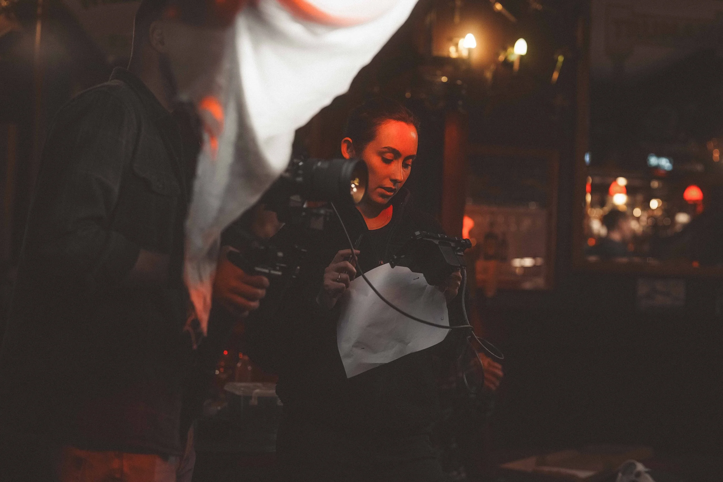 Two people, a man and a woman, are closely examining a computer screen in a dimly lit room. The woman holds a piece of paper. The room has warm lighting with lamps on the wall, and blurred lights are visible in the background.