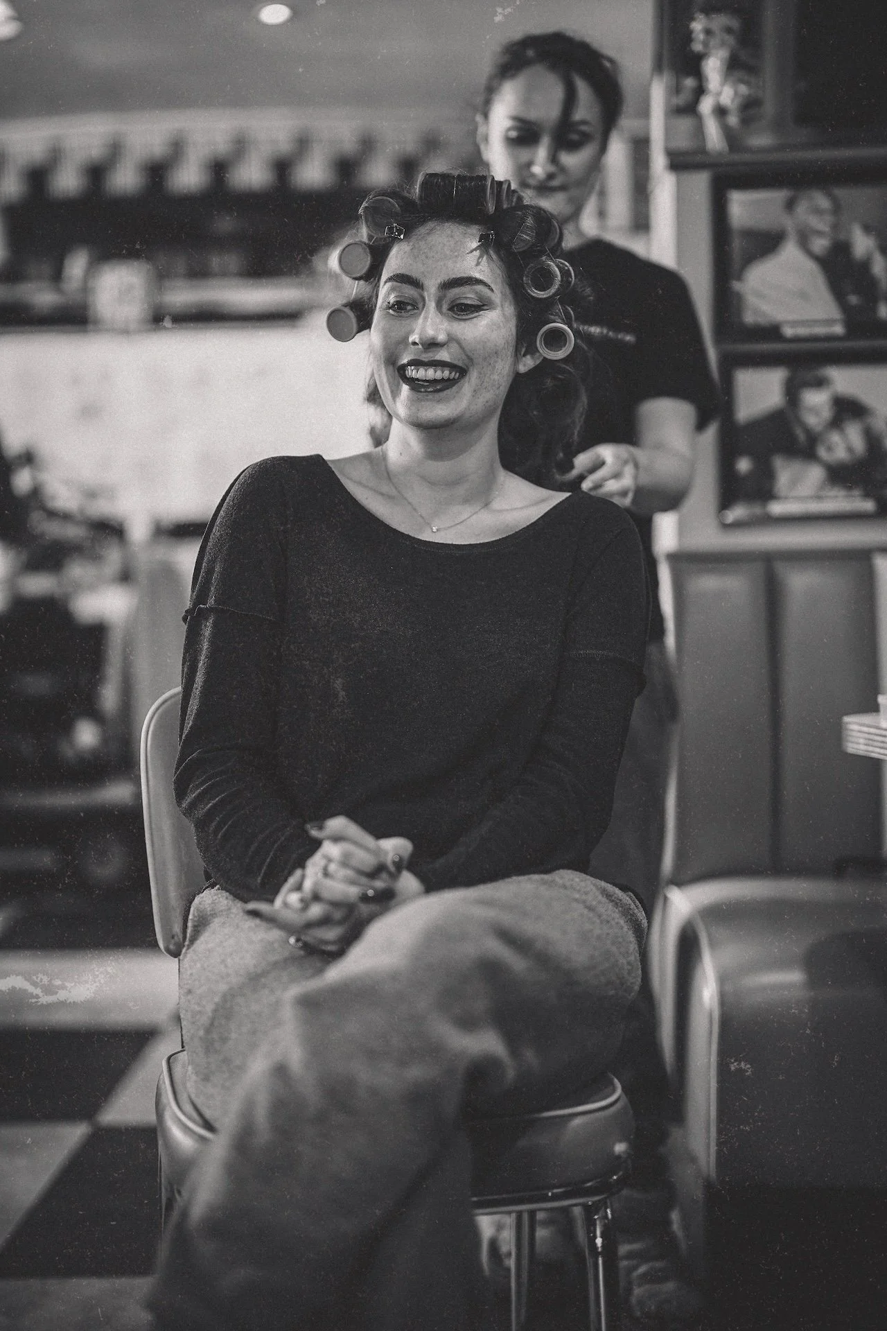 A woman with curlers in her hair sits smiling in a hair salon, with a stylist behind her. The salon has shelves with hair products and framed photos on the wall.
