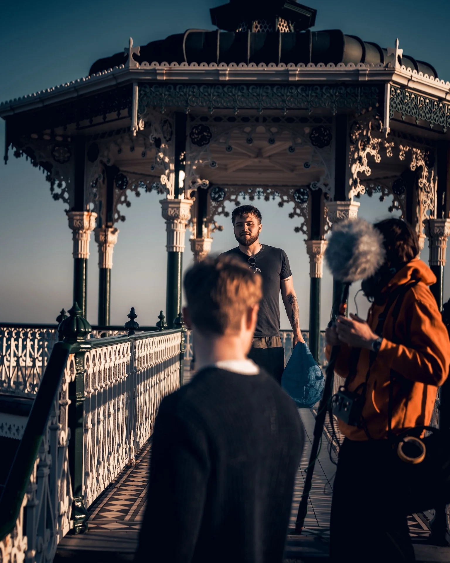 A man standing in front of a decorative pavilion on a rooftop, with people around, possibly filming or taking photos during sunset.