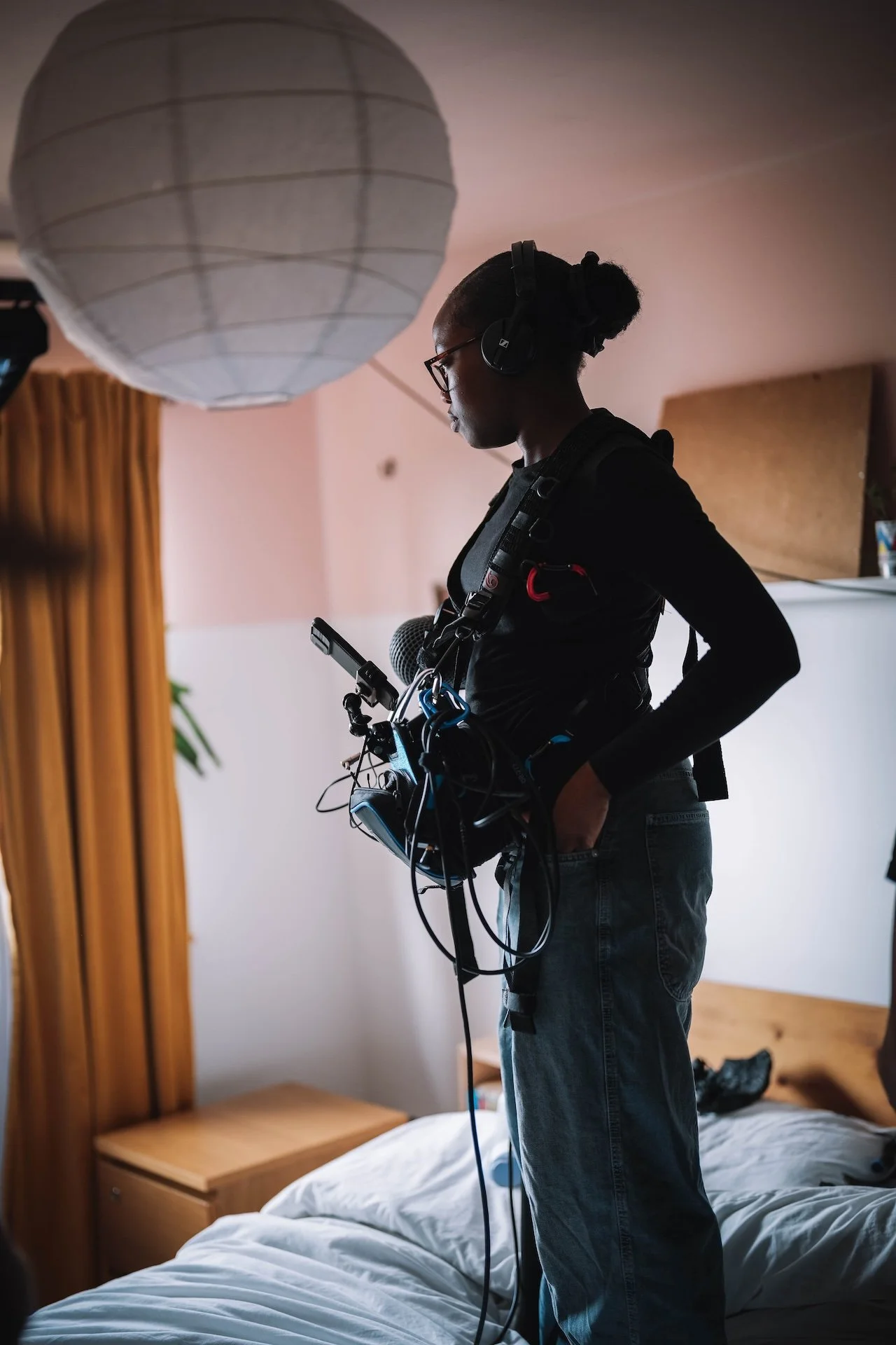 A person with glasses and headphones, holding equipment with wires, standing next to a bed in a bedroom with pink walls, a wooden headboard, and an orange curtain.
