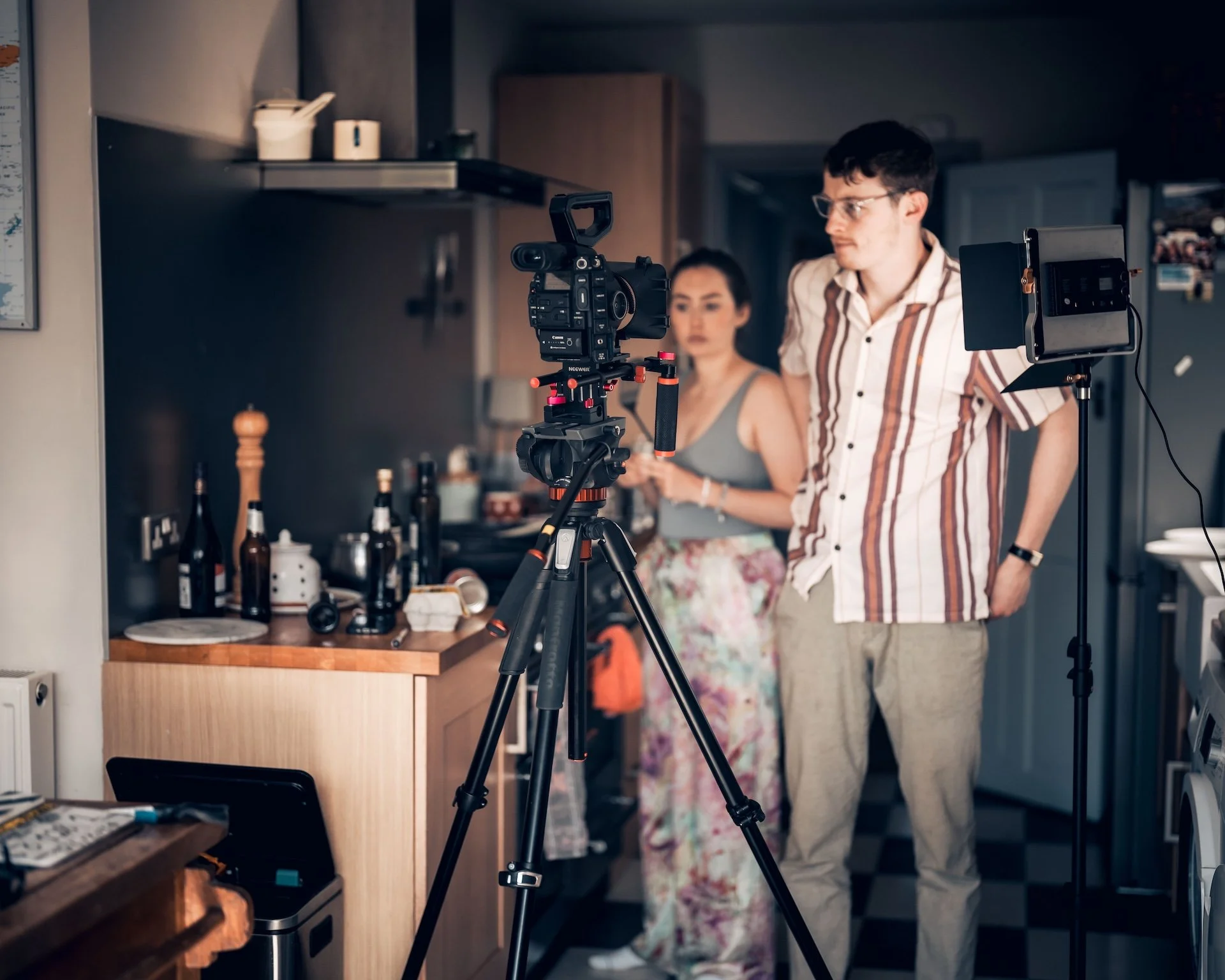Two people filming in a kitchen. A man in a striped shirt and glasses is standing near a camera on a tripod, and a woman in floral pants and a gray top is in the background. There are bottles and kitchen items on the counter.