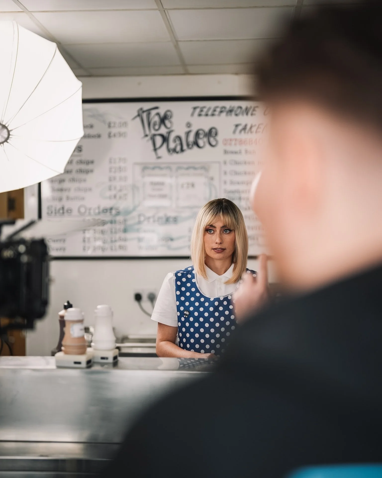A woman with blonde hair and blue eyes wearing a blue polka dot dress and white shirt standing in front of a menu board in a restaurant or café setting.