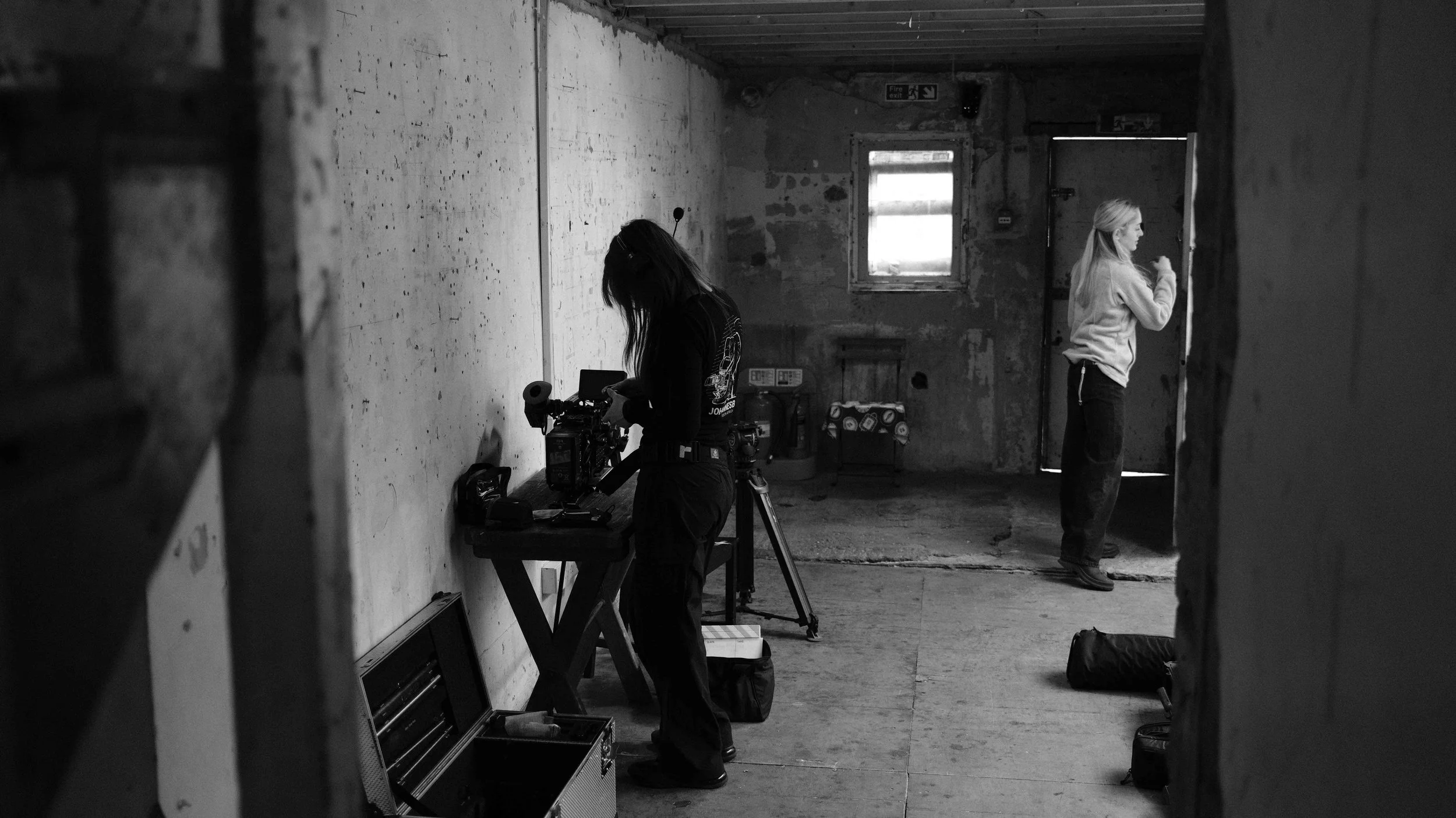 Black and white photo of two women in a rustic, unfinished room. One woman is working with film equipment on a table, while the other woman stands near a door, looking outside or at her phone.