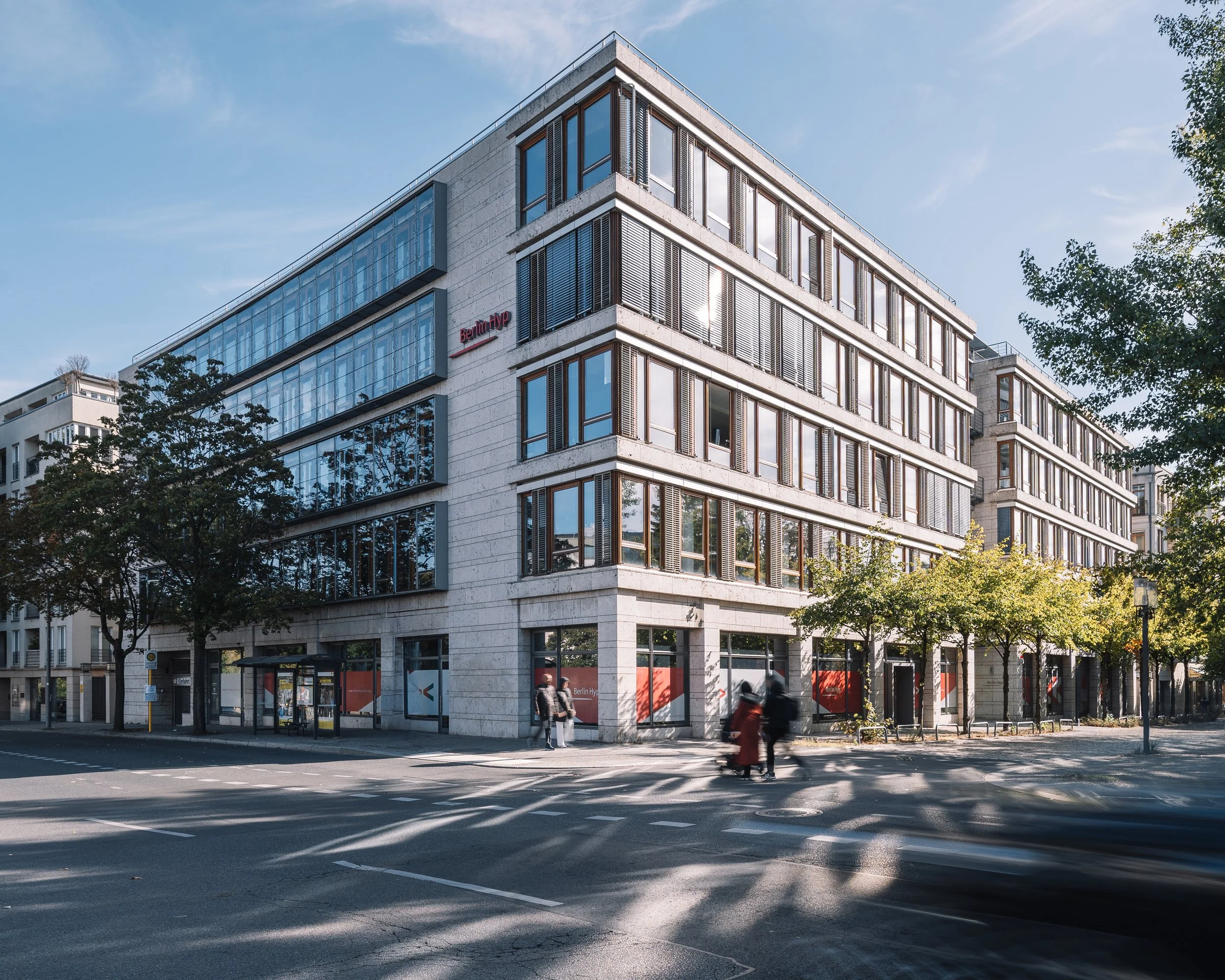 Modern multi-story office building with large glass windows on a city street, trees lining the sidewalk, and people walking and biking.