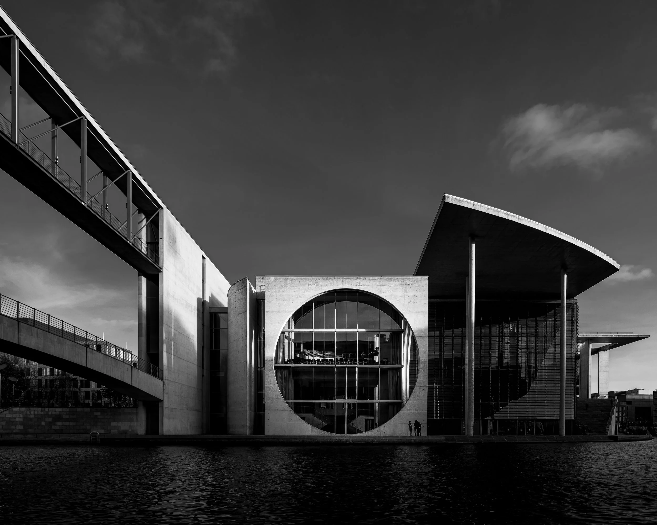 Modern architectural building with large circular window, flat overhanging roof supported by thin columns, and a bridge on the left side, reflected in water, in black and white.
