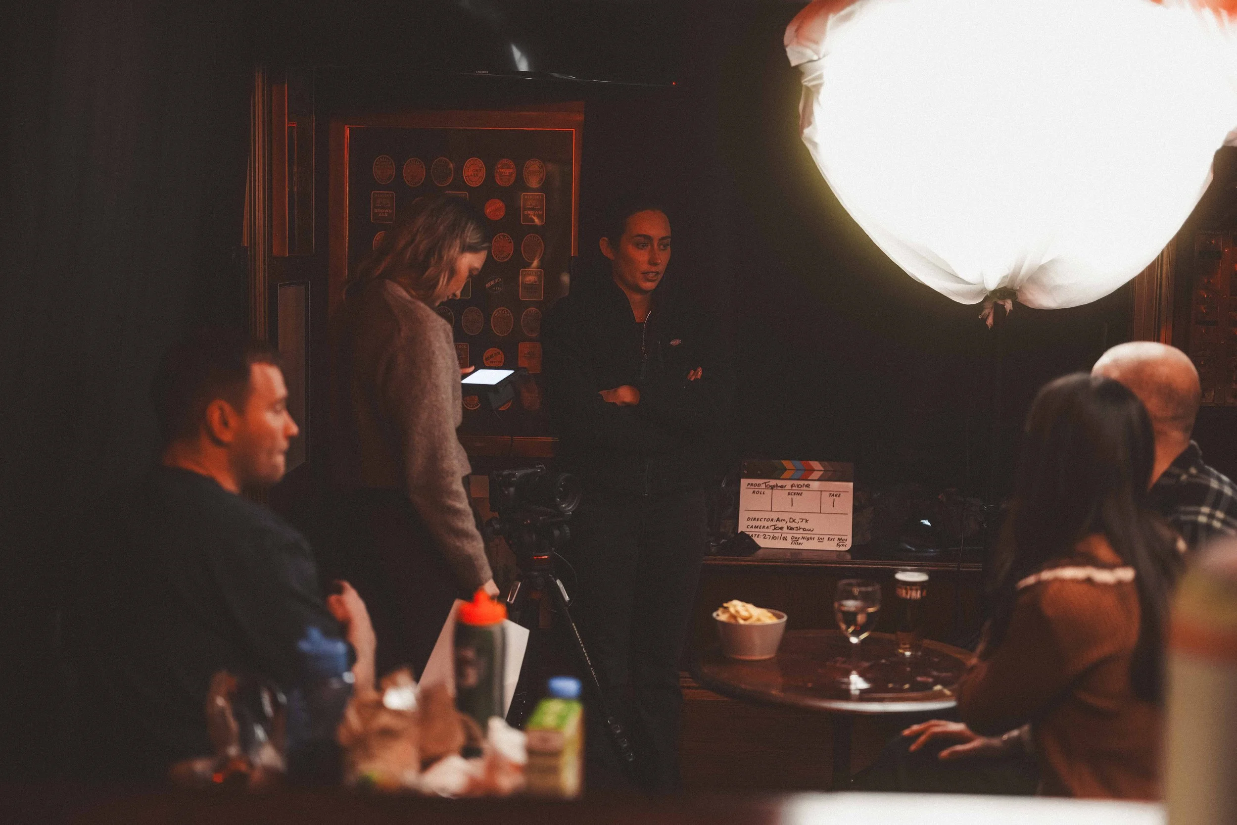 People sitting at tables in a dimly lit room with a large white balloon and a scene of a film or photo shoot setup, including a clapperboard and film crew.