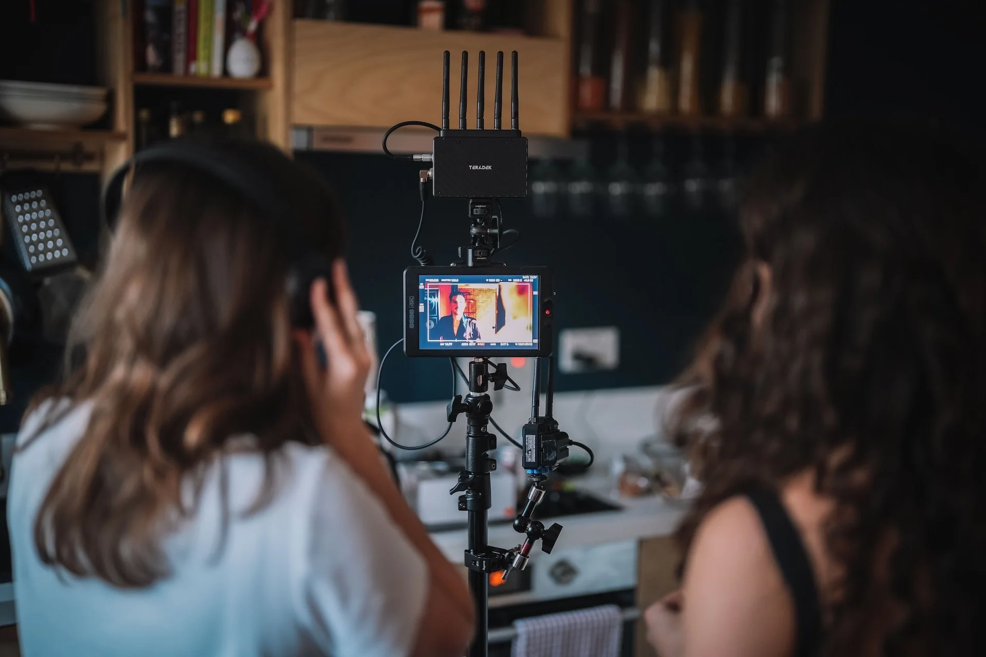Two women working with film equipment on a film set, with a camera filming a woman standing against a wall. The camera operator is wearing headphones and looking at the camera while controlling it, and the woman on set is speaking or acting.