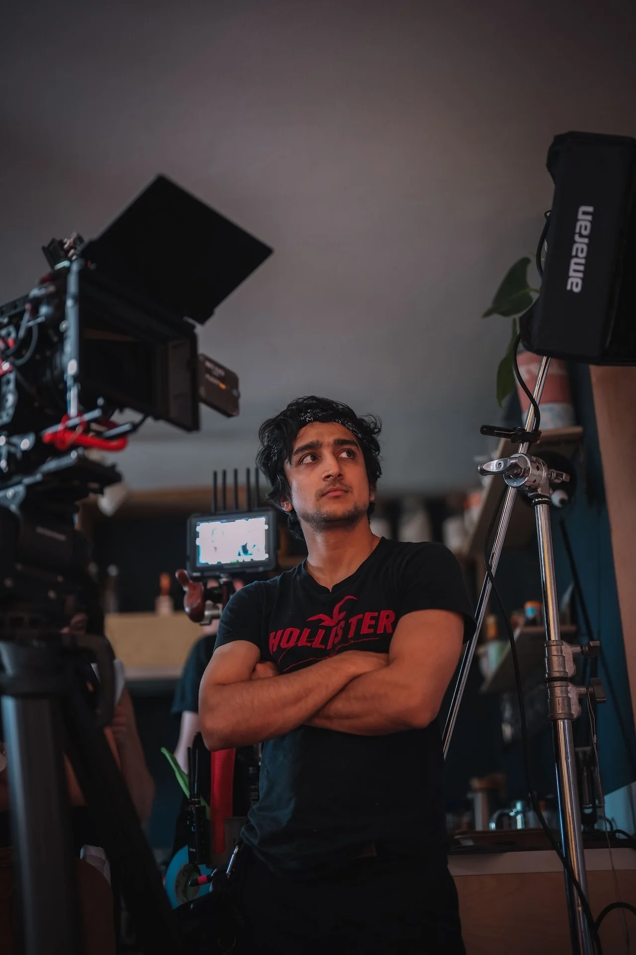 Young man with arms crossed standing in front of film equipment on a film set, behind others working, indoors with ceiling and shelves.