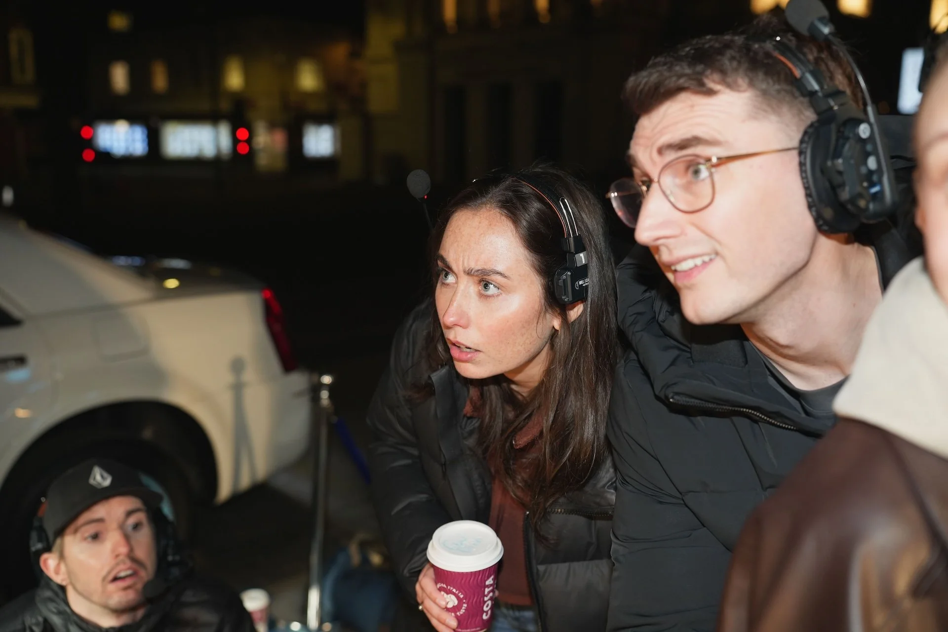 Three people wearing headsets are closely looking at something off-camera. One woman in the middle holds a coffee cup, and a man at the bottom left wears a helmet with headphones, seemingly inside a city street at night.