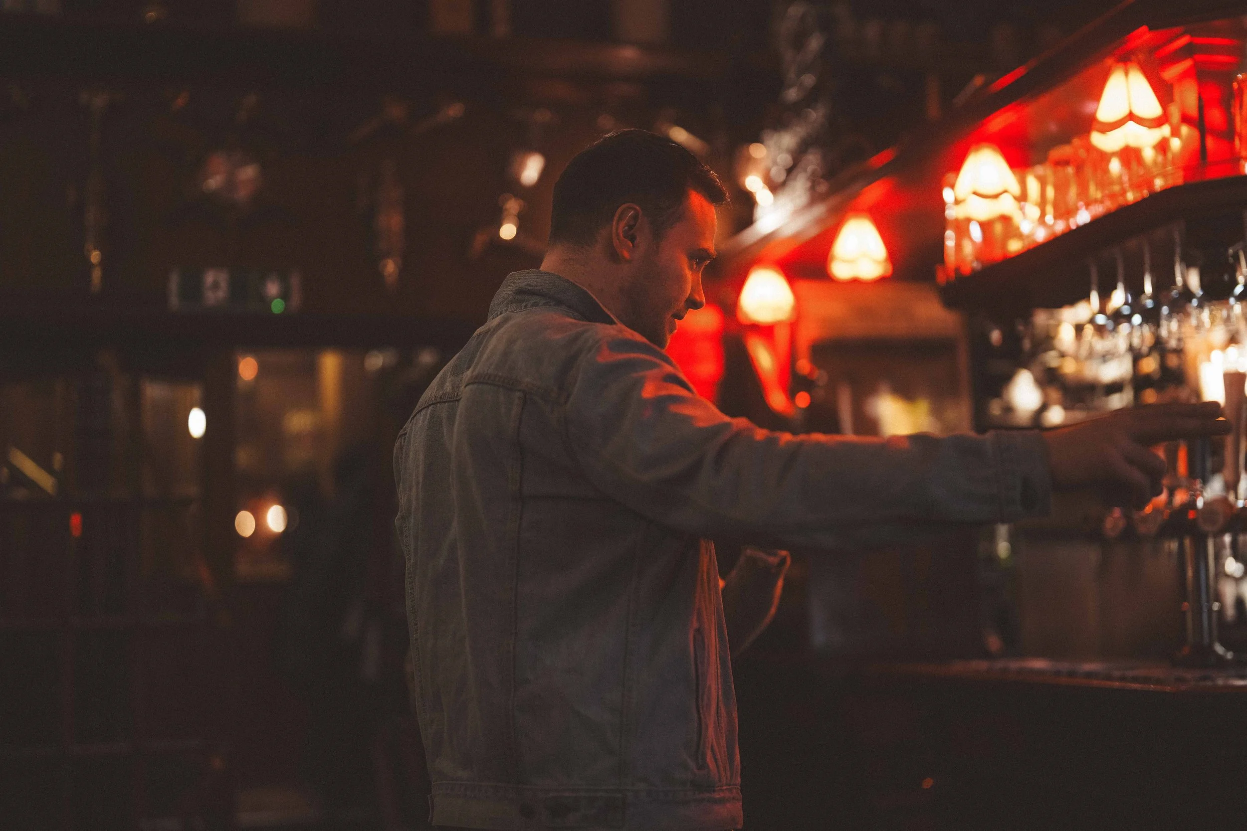 A man wearing a gray jacket with short dark hair standing in a dimly lit bar with red lamps on the wall and a shelf of liquor bottles in the background.
