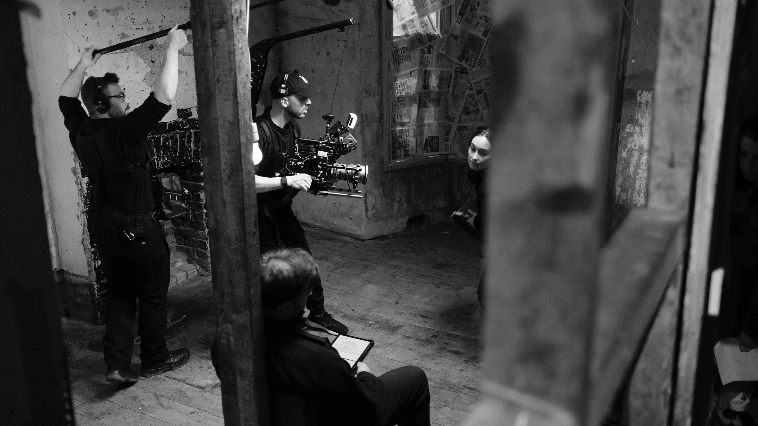 A black-and-white photo of a film shoot behind the scenes, with a cameraman holding a camera rig, two crew members adjusting equipment, and a woman sitting in front of a wall covered with newspapers or posters.