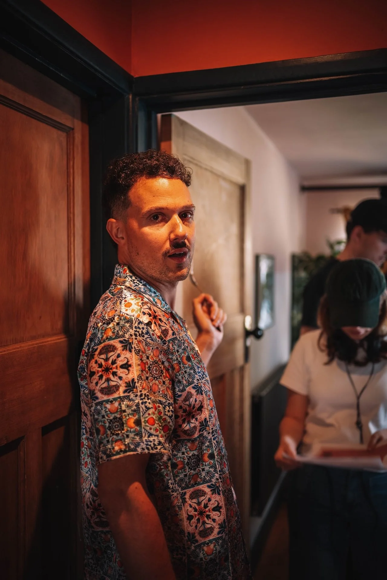 A man with curly hair and a mustache wearing a colorful patterned short-sleeve shirt is standing in a room with wooden walls and a black frame around the doorway. He is looking toward the camera. In the background, a woman with long dark hair and a w