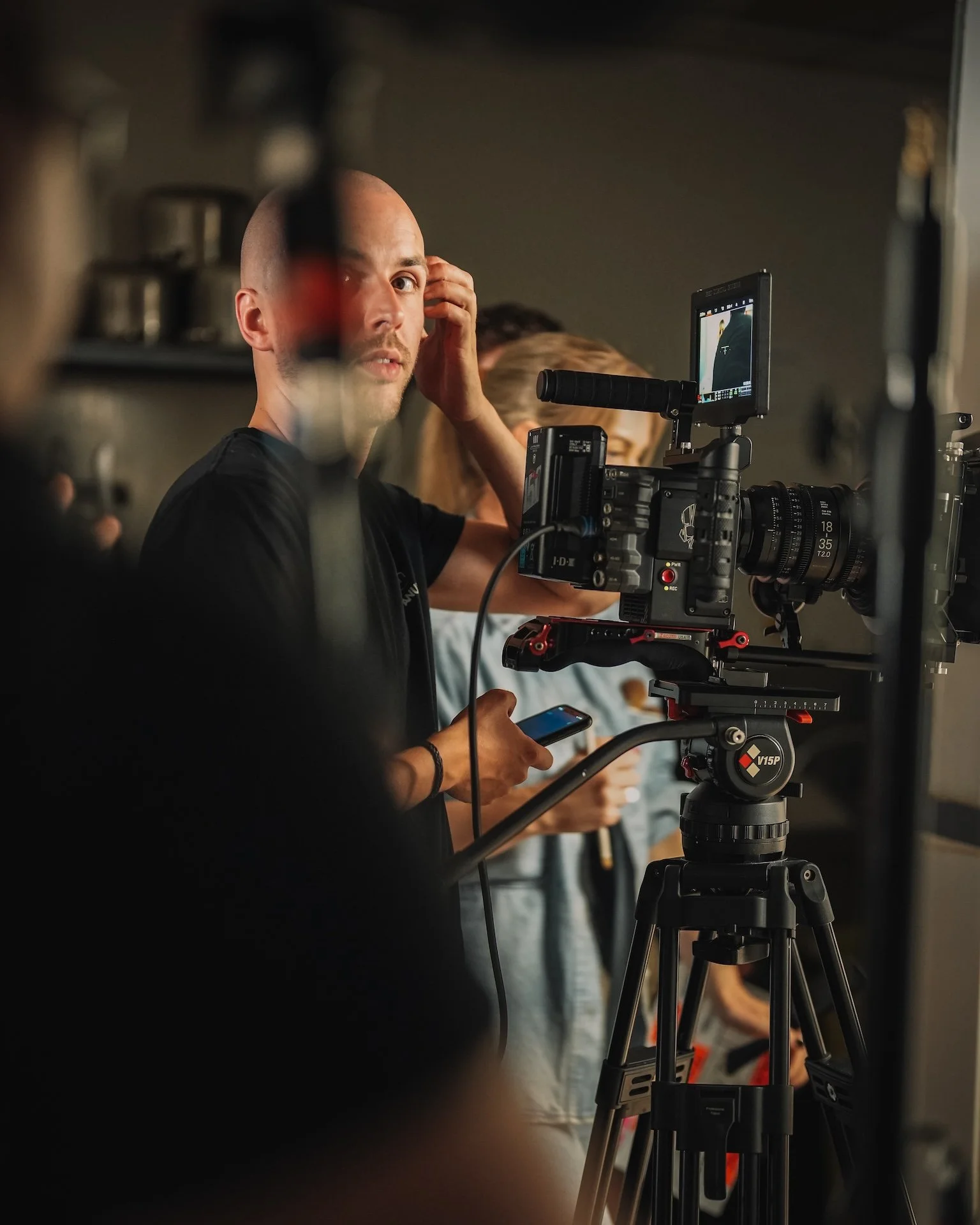 A man with a shaved head adjusts his ear while operating a professional camera on a tripod, with other crew members in the background.