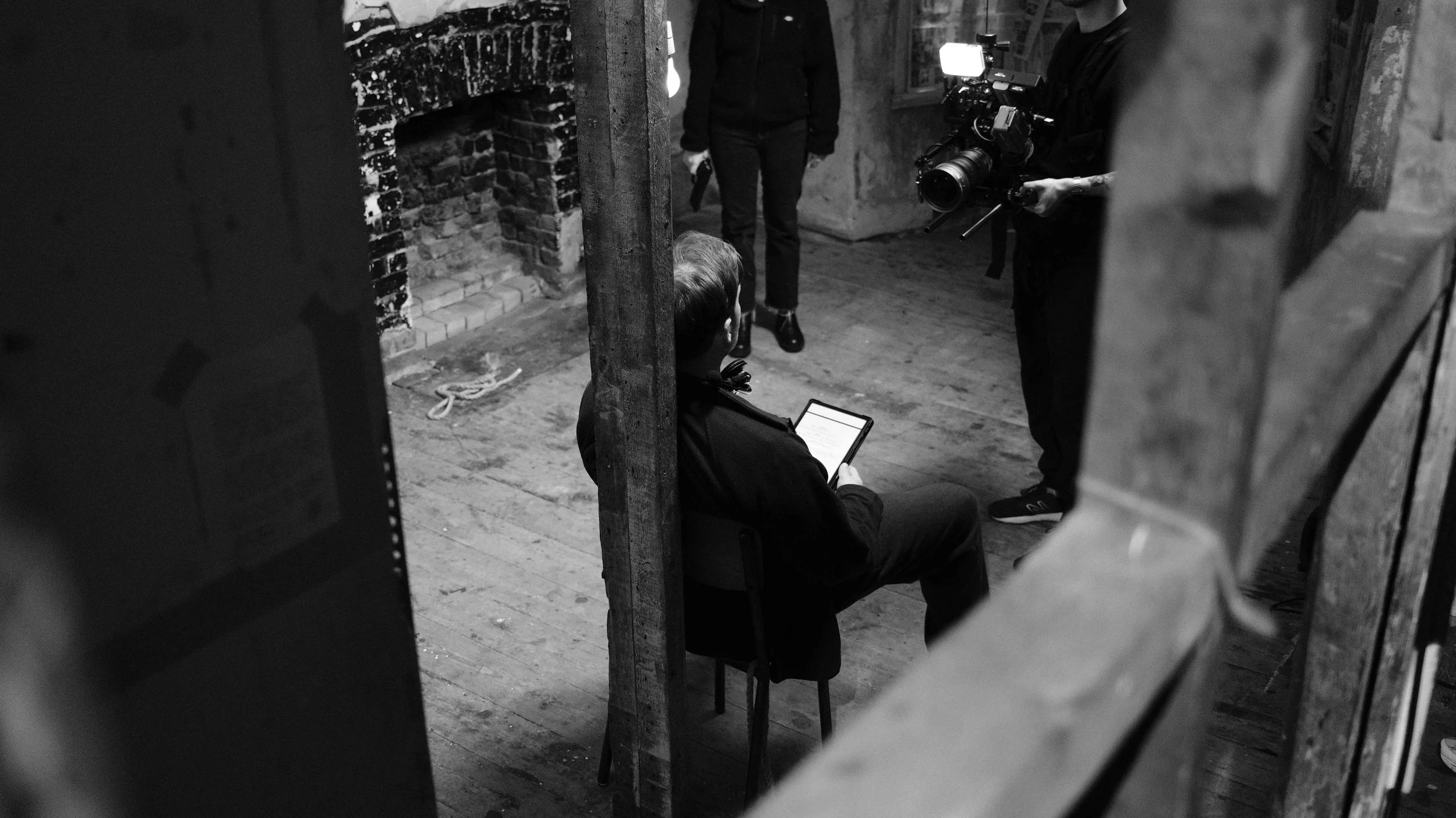 Black and white photo of a person sitting and looking at a tablet, while a camera operator records them, in a rustic room with brick fireplace and wooden floors.