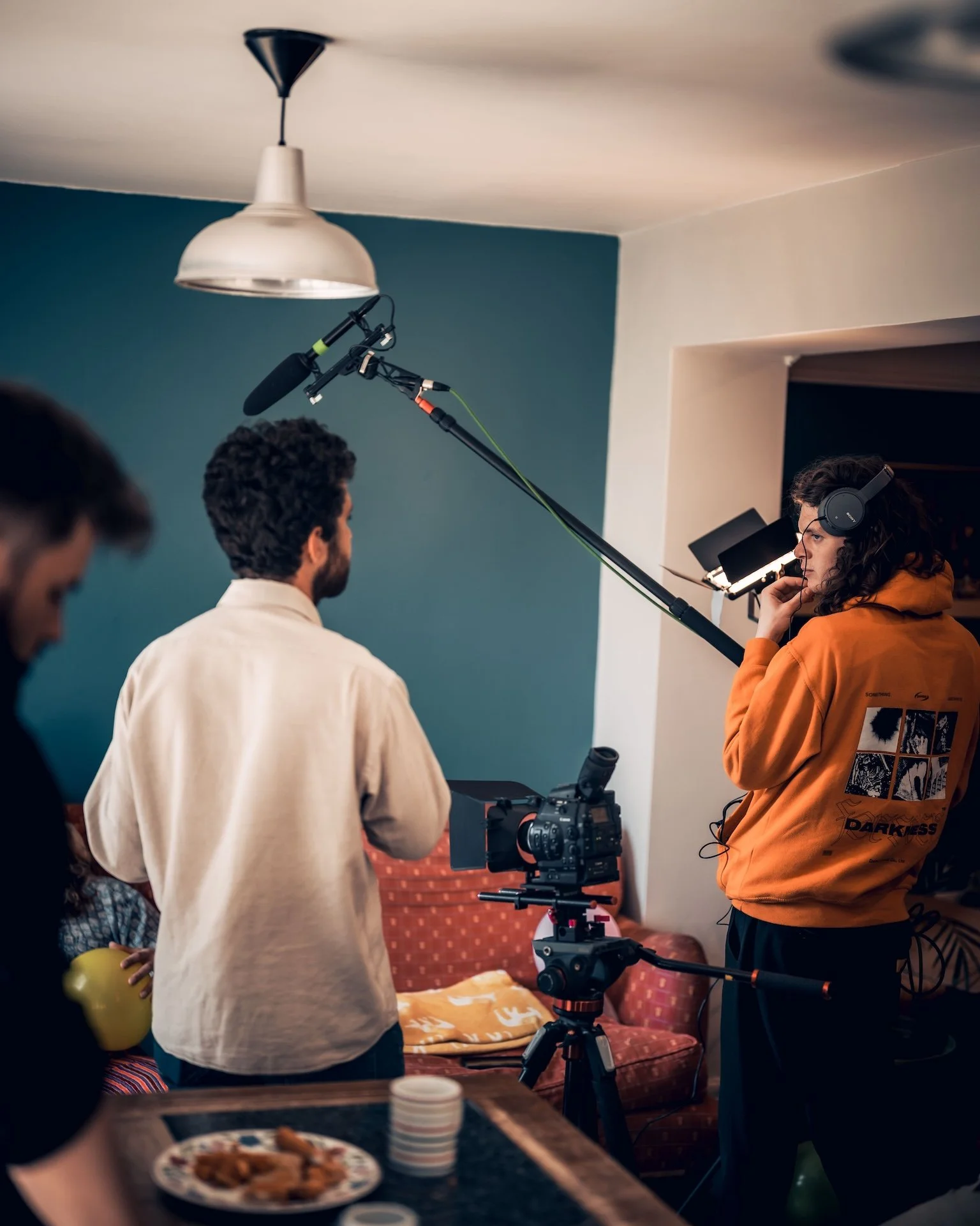 Filmmakers filming a scene indoors with camera equipment, a man in a beige shirt, and a woman in an orange hoodie wearing headphones.