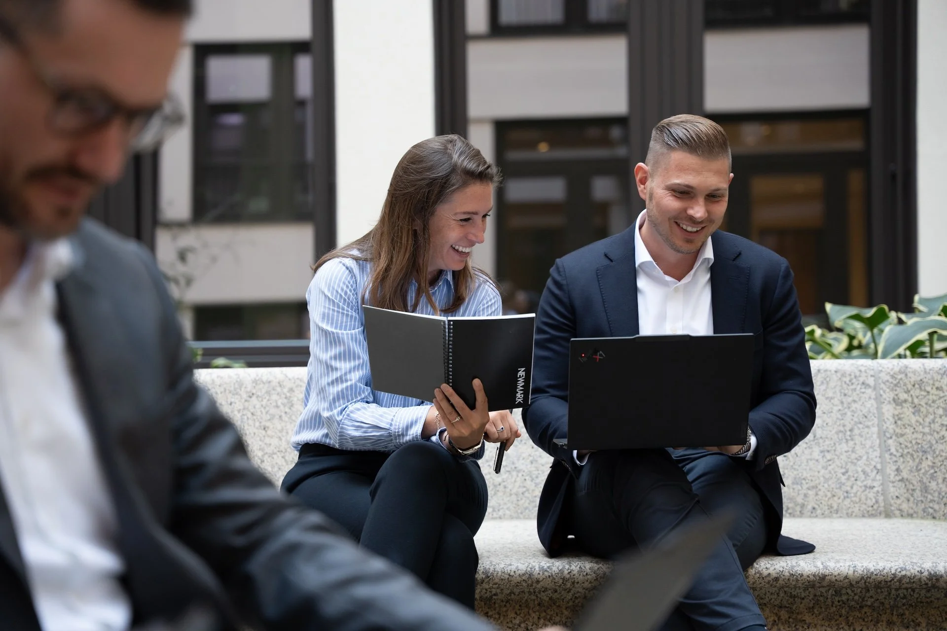 Two people sitting on a bench, looking at a laptop and sharing a laugh, in a professional outdoor setting.