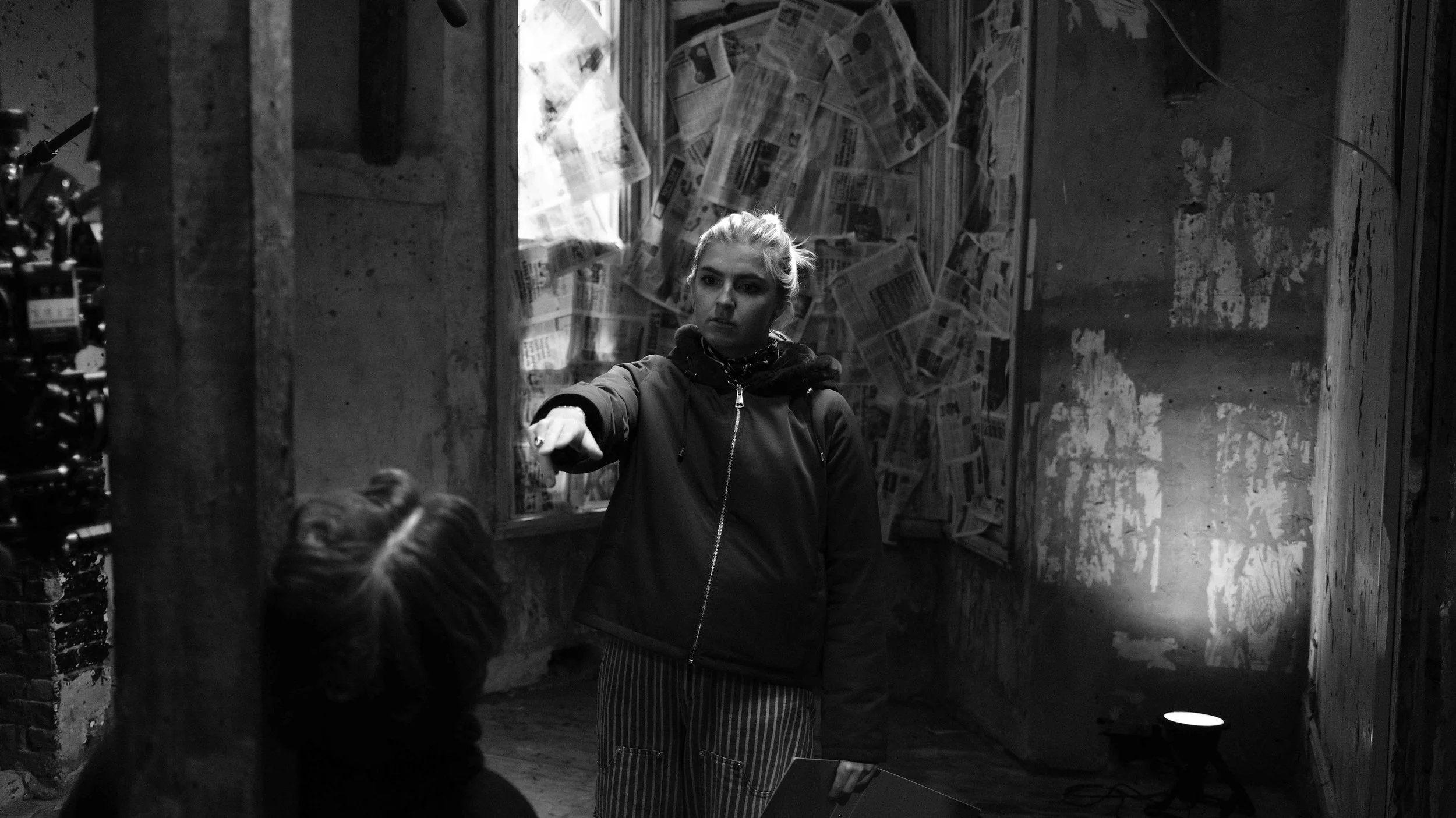 A woman with blonde hair standing indoors, pointing her finger at someone who is seated and viewed from behind. The room has walls with peeling paint and a background covered with newspapers. There is a small spotlight on the floor illuminating the s