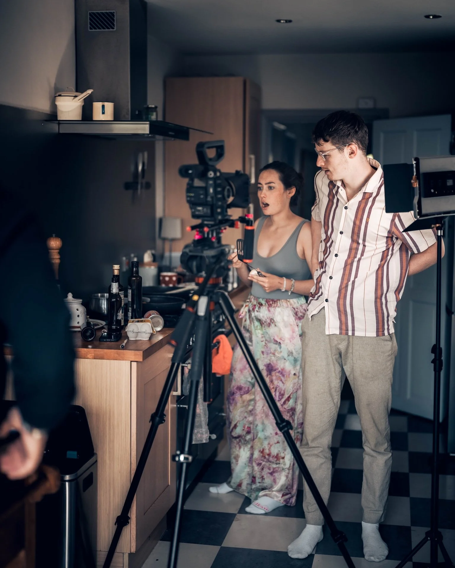 Two people filming a scene in a kitchen. The woman has dark hair, wears a gray top and floral pants, and looks surprised. The man has dark hair, glasses, wears a striped shirt and beige pants. Camera equipment is set up around them.