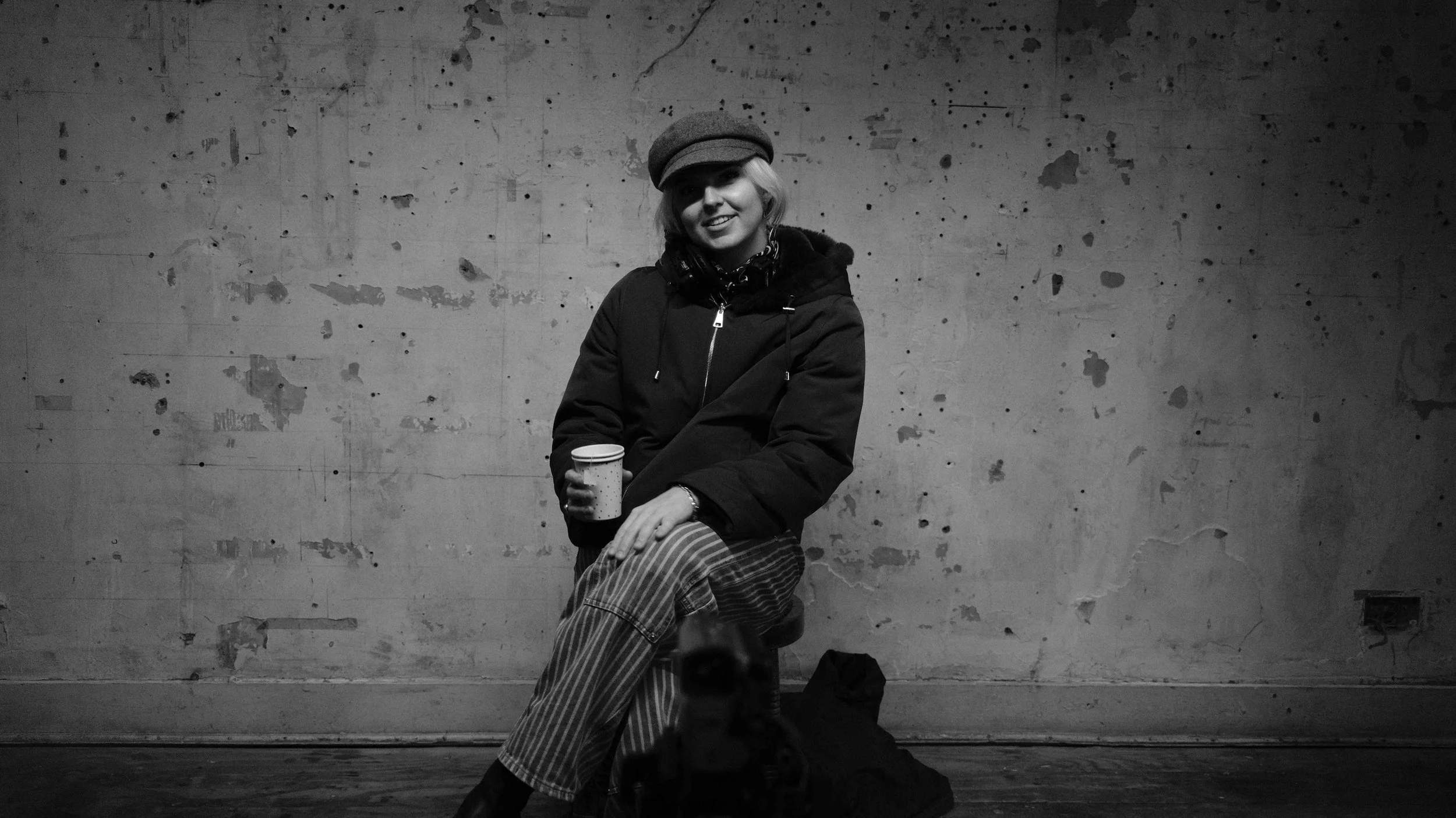 A woman sitting on a stool against a grungy, textured wall, holding a disposable coffee cup, wearing a hat, scarf, and striped pants, smiling at the camera in a black and white photo.