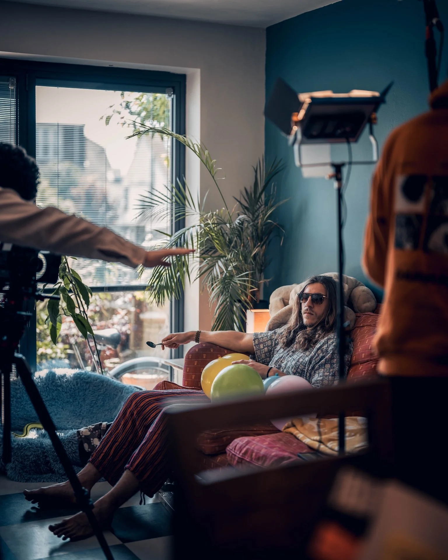 A person with long hair and sunglasses sitting on a couch, holding balloons and a spoon, in a room with large window and indoor plants, while behind a film crew is filming.