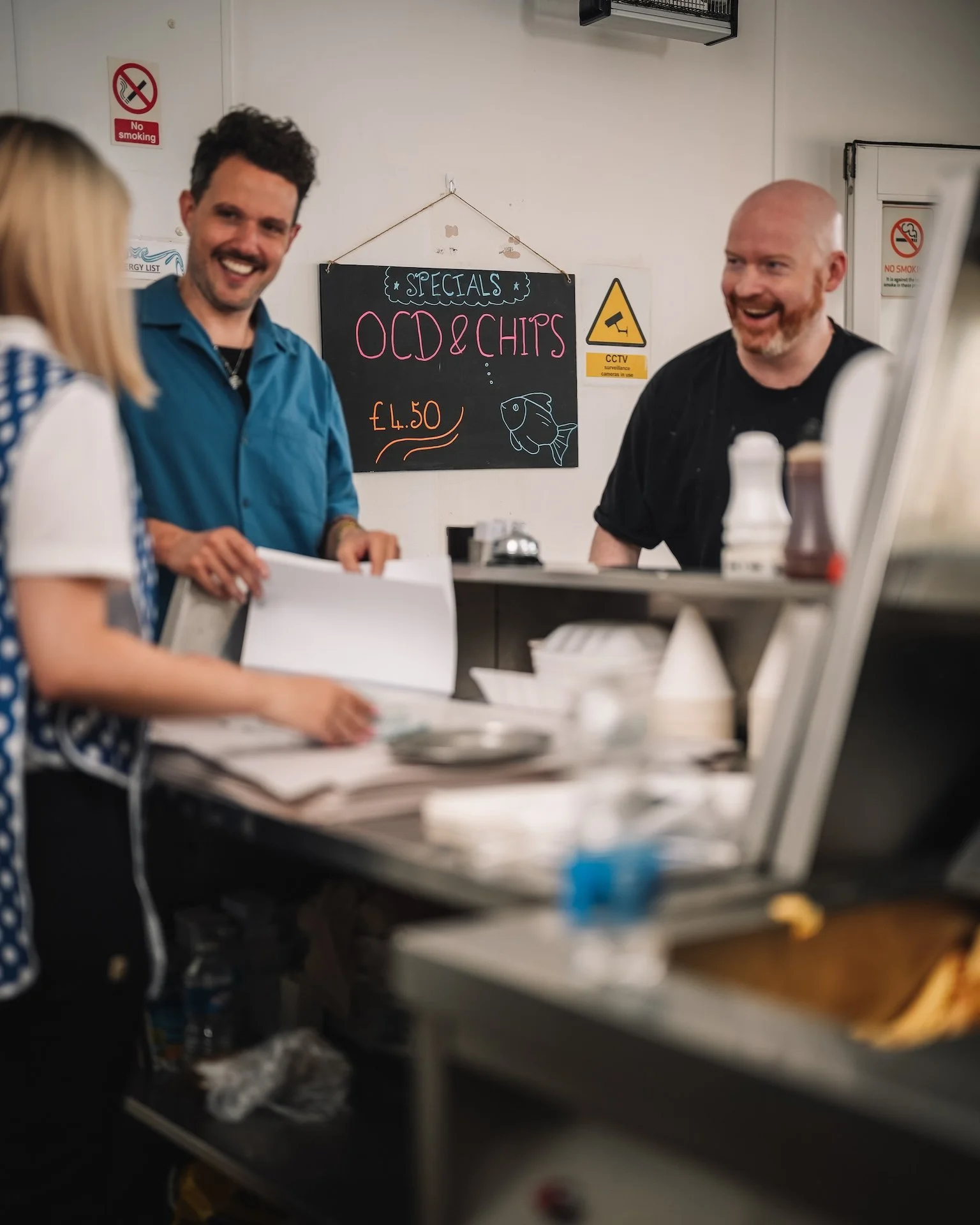 Two men are smiling and conversing inside a restaurant or cafe, with a woman and a chalkboard sign behind them displaying specials including 'OCD & Chips' priced at £4.50. The background includes a CCTV warning sign and a no smoking sign.