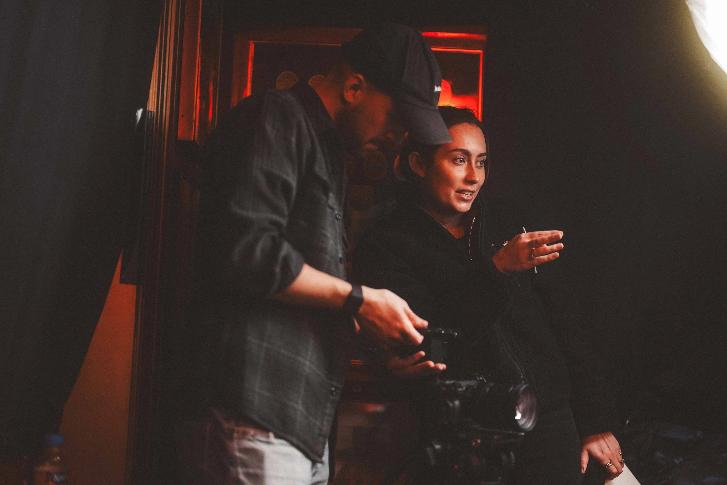 Two people in a dimly lit room, one wearing a cap and checking something on a device, the other smiling and holding a cigarette.