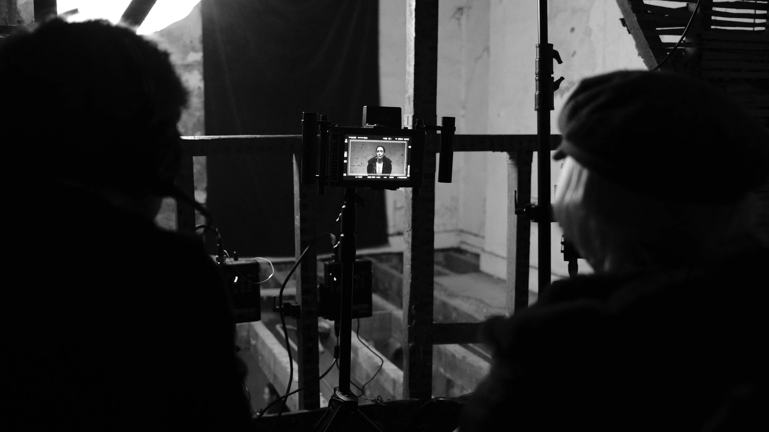 Two people on a film set behind the camera, one with dark hair on the left and one wearing a beanie on the right, with a monitor showing a woman in a blazer being recorded.