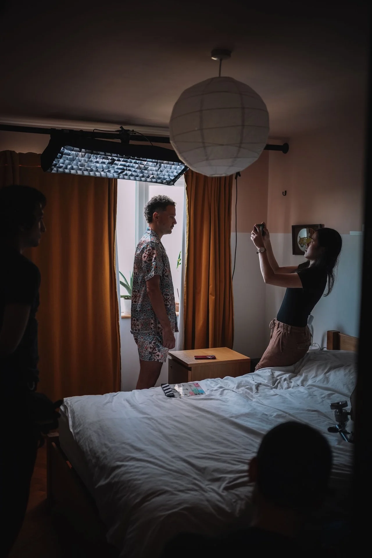 A person standing on a bed with a white sheet, surrounded by others in a dimly lit room with a window, orange curtains, and a white paper lantern hanging from the ceiling. A woman is taking a photo of the person on the bed.