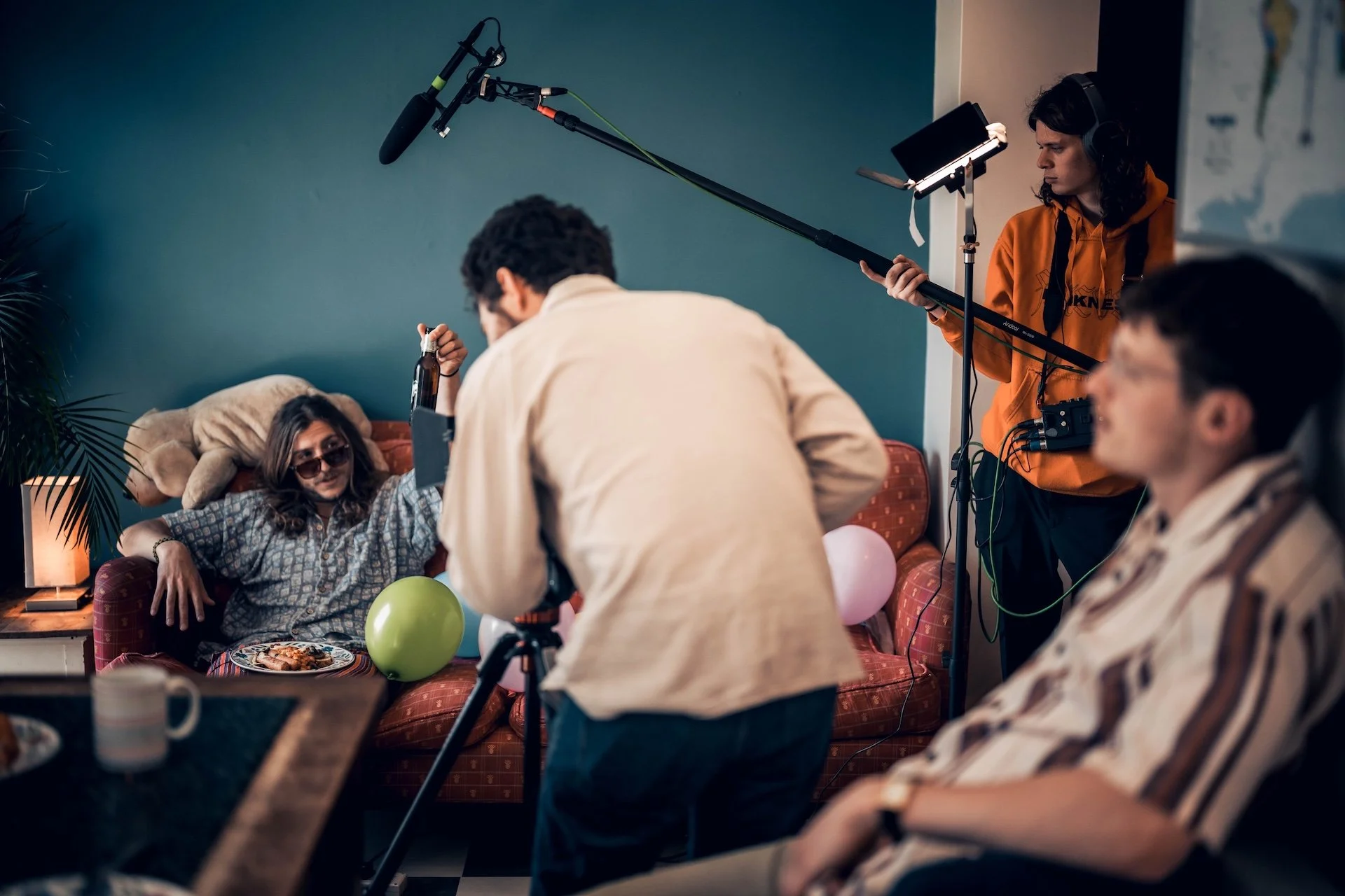A behind-the-scenes look at a video production set, featuring a group of young people in a cozy living room with balloons, snacks, and drinks, while a crew member with professional audio equipment films them.
