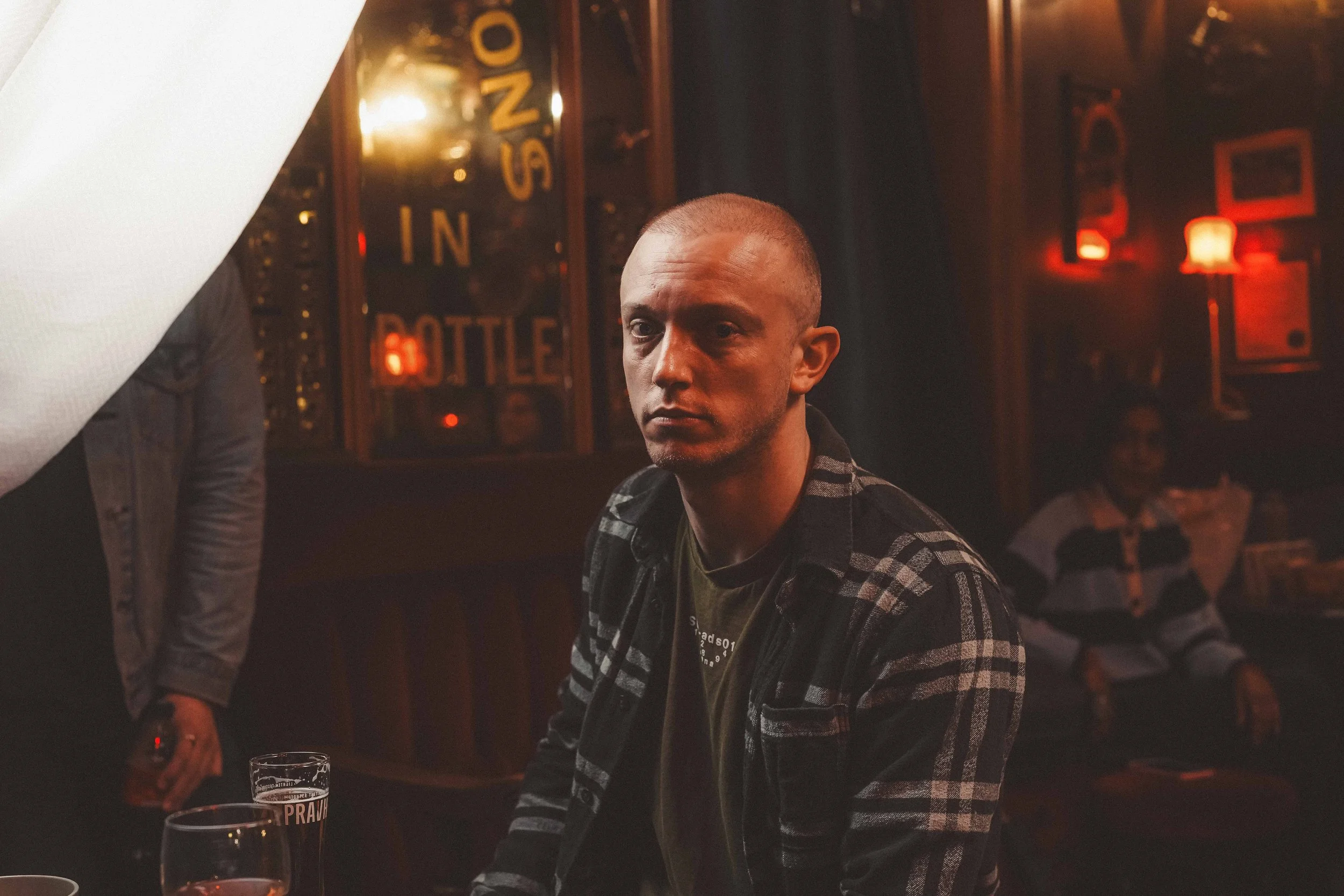 A man with a buzz cut hairstyle wearing a black and white checkered shirt in a dimly lit bar or pub, with reddish-orange lighting and framed pictures on the wall in the background. There are glasses on the table in front of him, one labeled 'PRANK'.