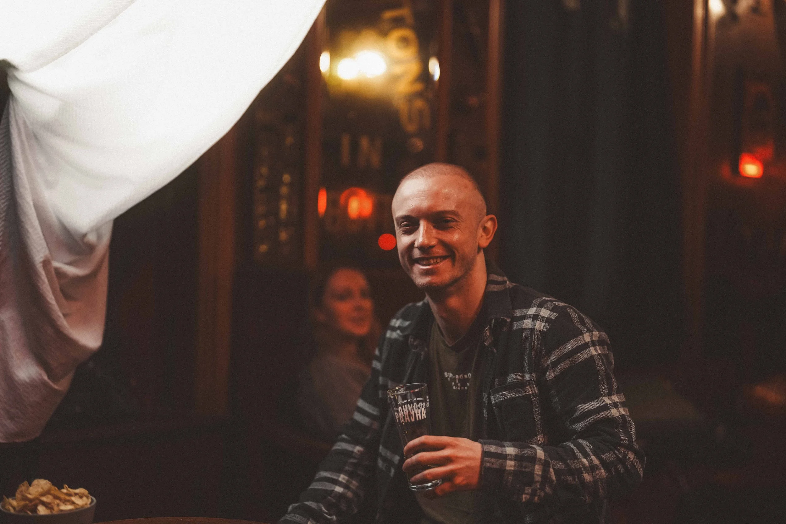 Smiling man holding a glass of beer in a bar or restaurant with warm lighting and people in the background.