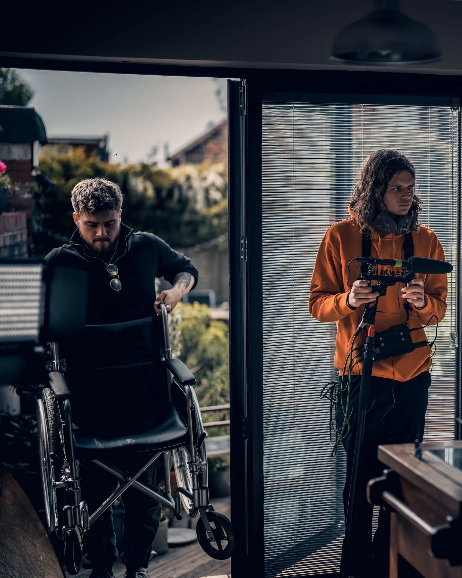 Two young adults, one with a disability, are engaged in filming or photography outdoors on a balcony or rooftop. The person on the right is holding camera equipment, while the person on the left appears to be managing another piece of equipment or a 