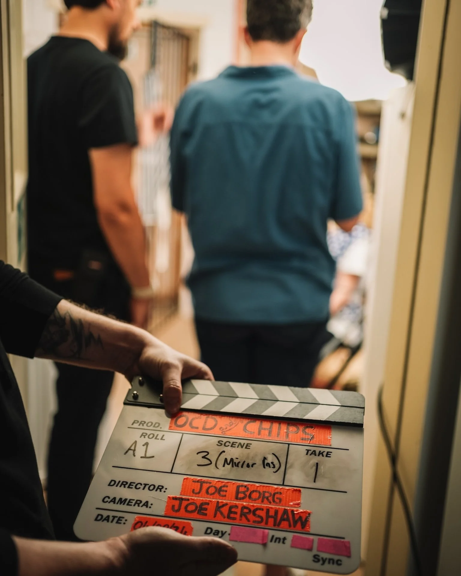 Close-up of a film director or crew member holding a clapperboard with handwritten details. In the background, appears to be two people in conversation or scene, slightly blurred, inside a room or hallway.