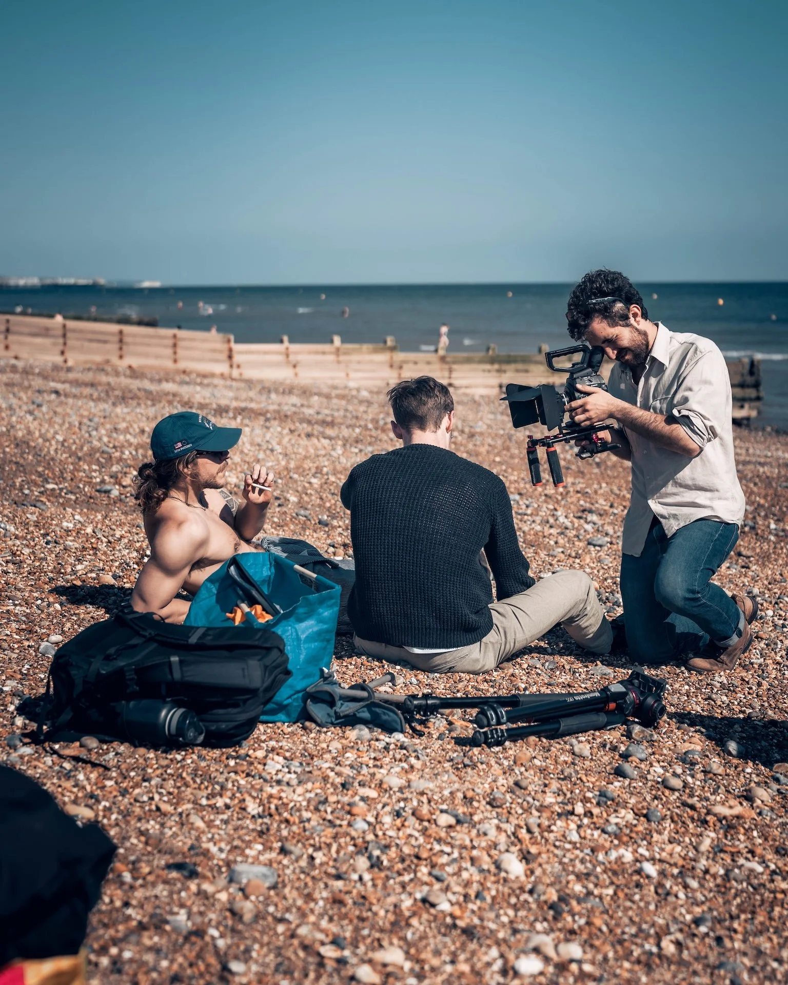 Three men are sitting on a pebbled beach by the ocean, filming a scene. One man is shirtless, wearing a baseball cap, sunglasses, and smoking. Another man is wearing a black sweater, and the third man is operating a camera, standing and smiling. Ther