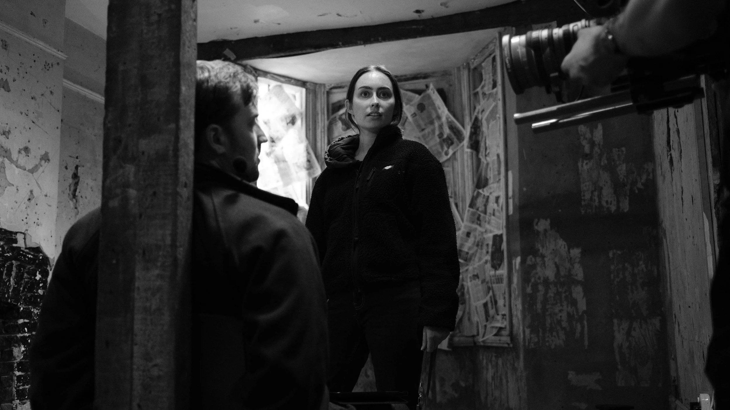 A black-and-white photo of a woman standing in a dimly lit, dilapidated room with newspapers taped on the wall behind her. She is looking slightly to the side and holding a bag. Two men are partially visible, one on the left side with his face turned